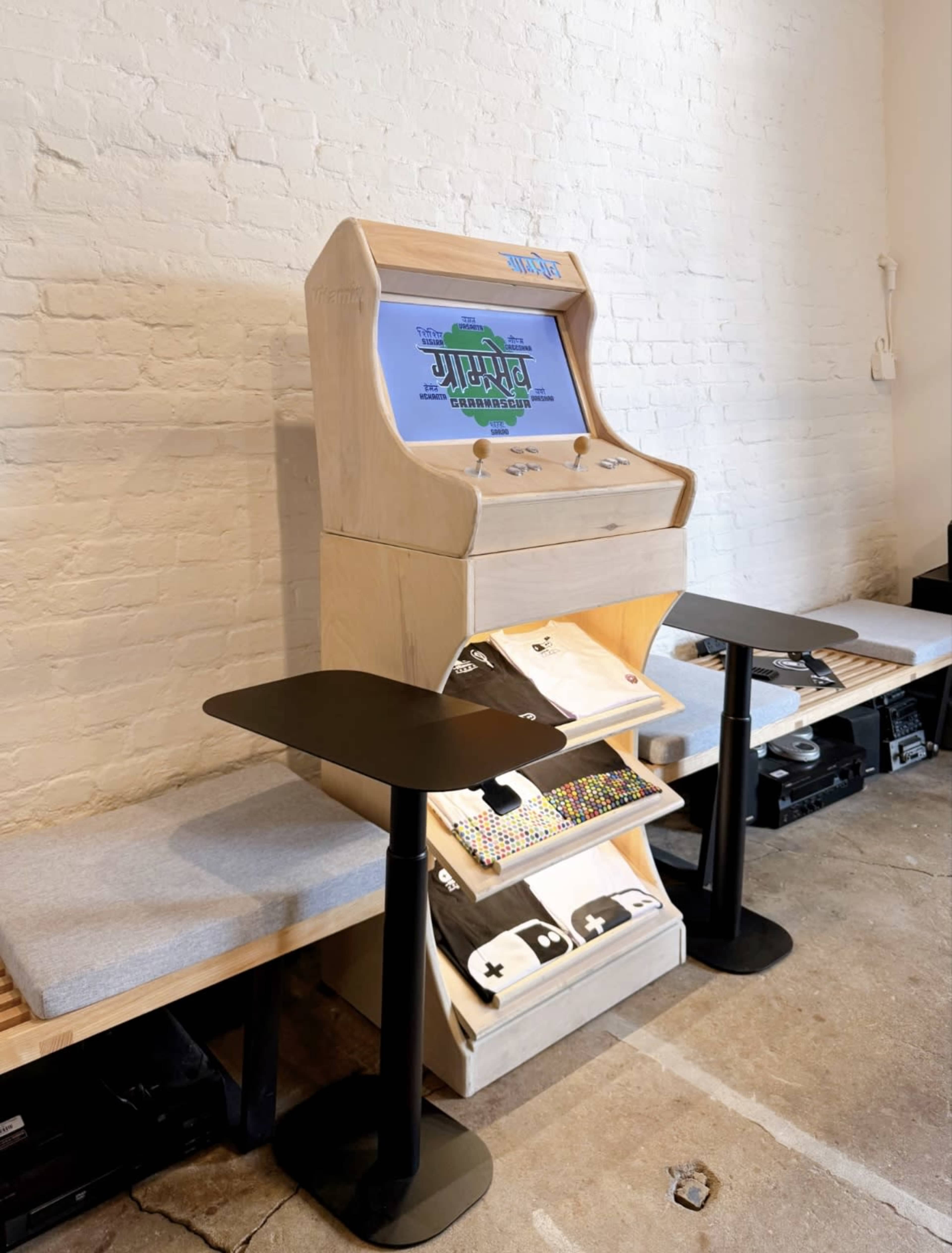 The image shows a wooden arcade game cabinet with a screen displaying a game, accompanied by two seating areas and small black tables.