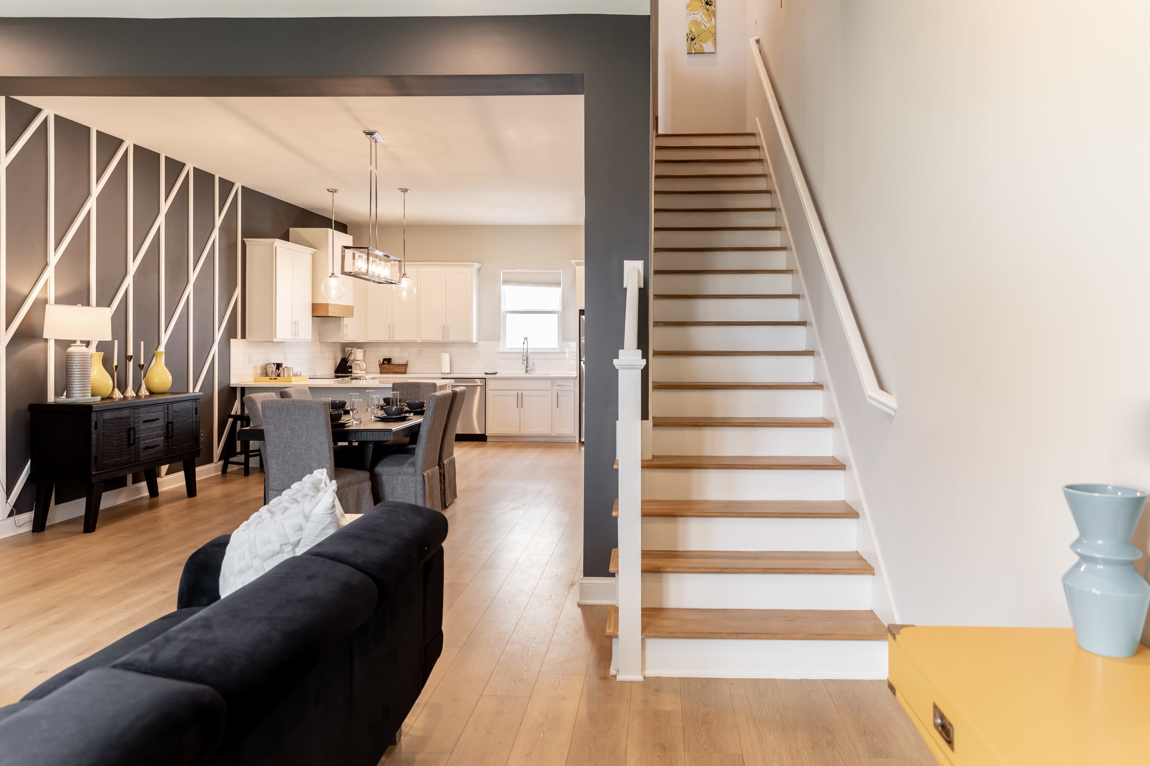 The image shows a modern interior with a staircase leading to an upper level, adjacent to a dining area featuring a black table and a patterned wall.
