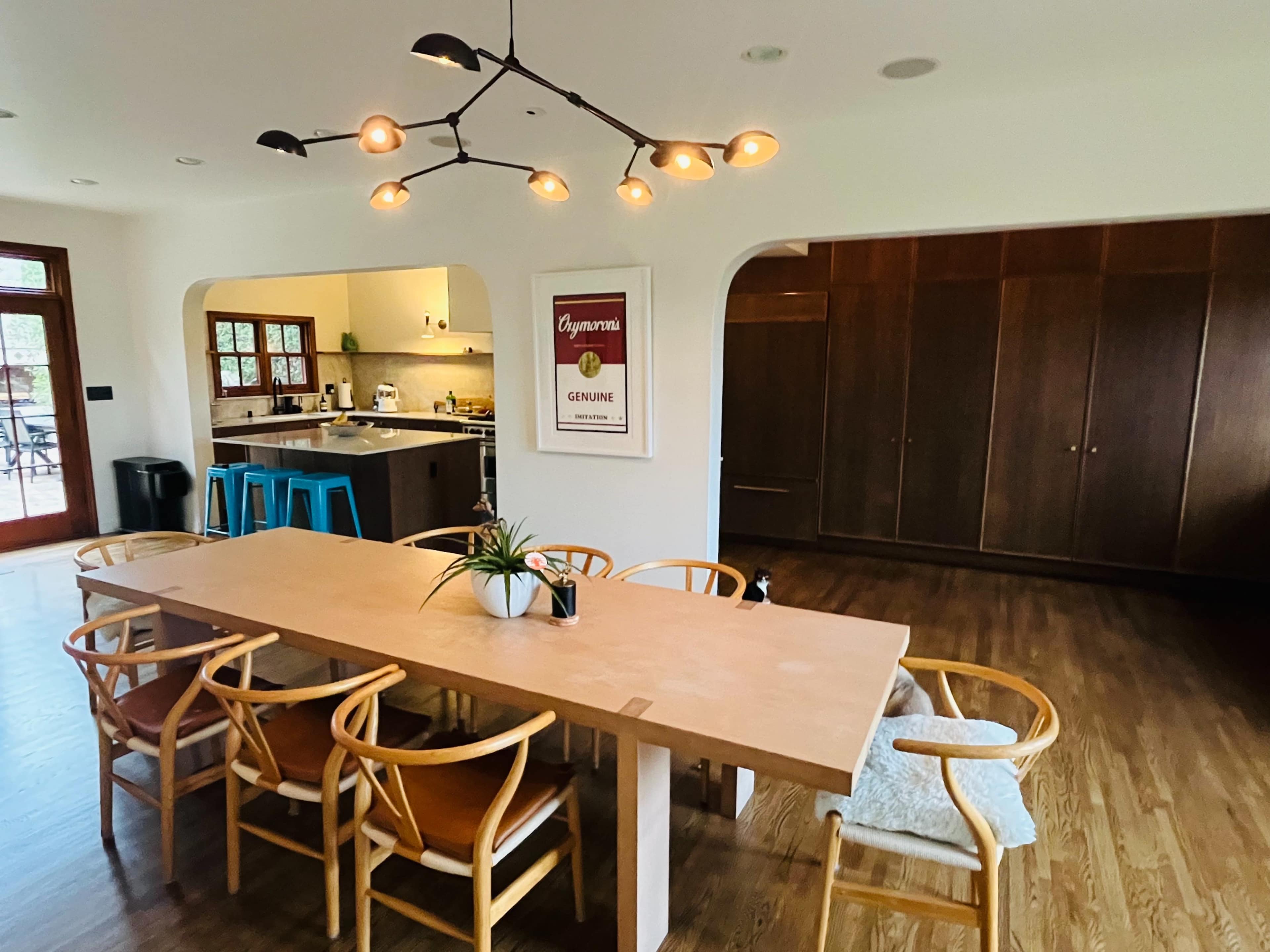 A modern dining area features a long wooden table surrounded by chairs, with a large arch leading to a kitchen and a wall of wooden cabinets in the background.
