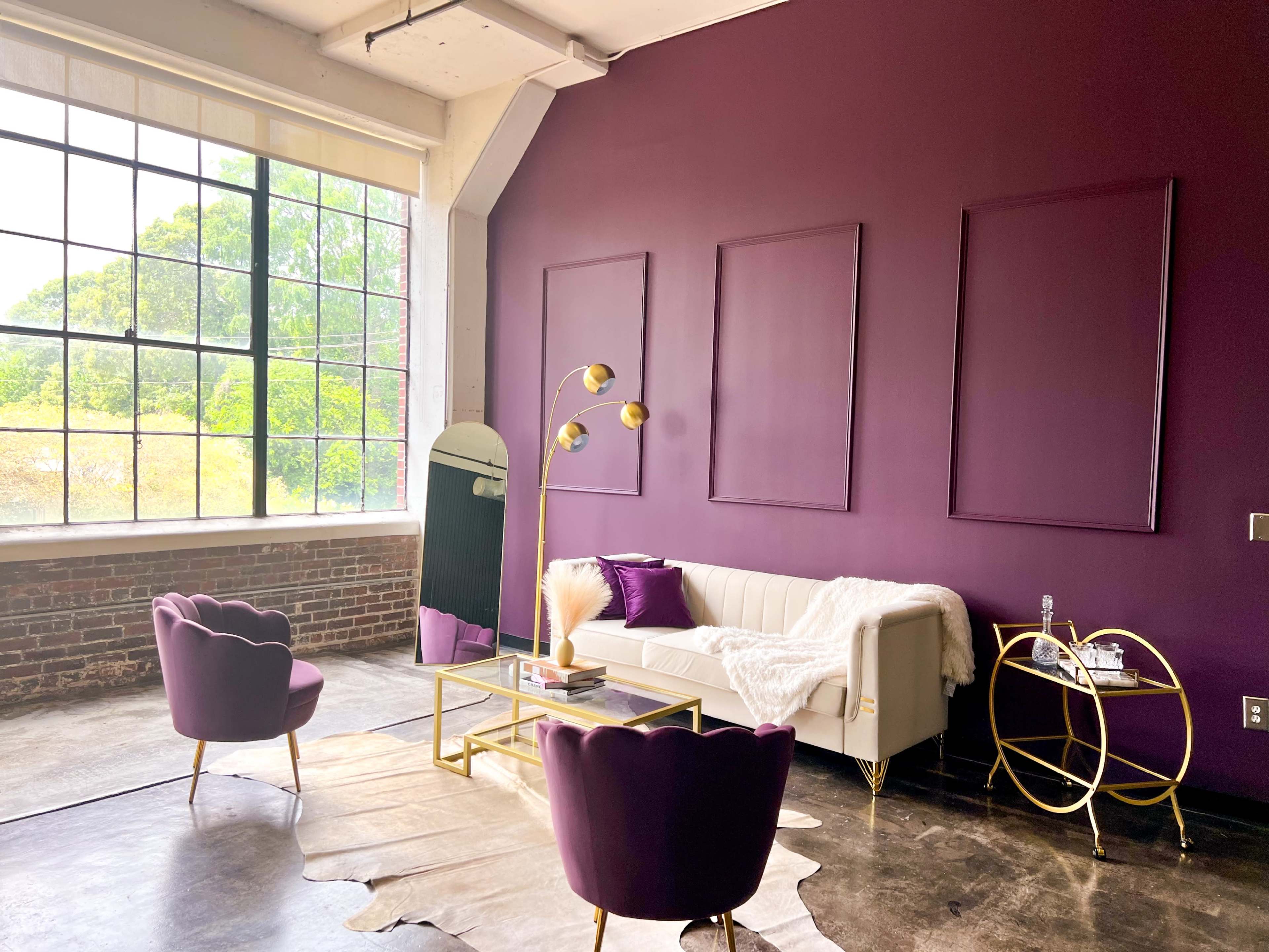 A modern living room features a white sofa, two purple chairs, a gold coffee table, and a large window with a view of greenery outside.
