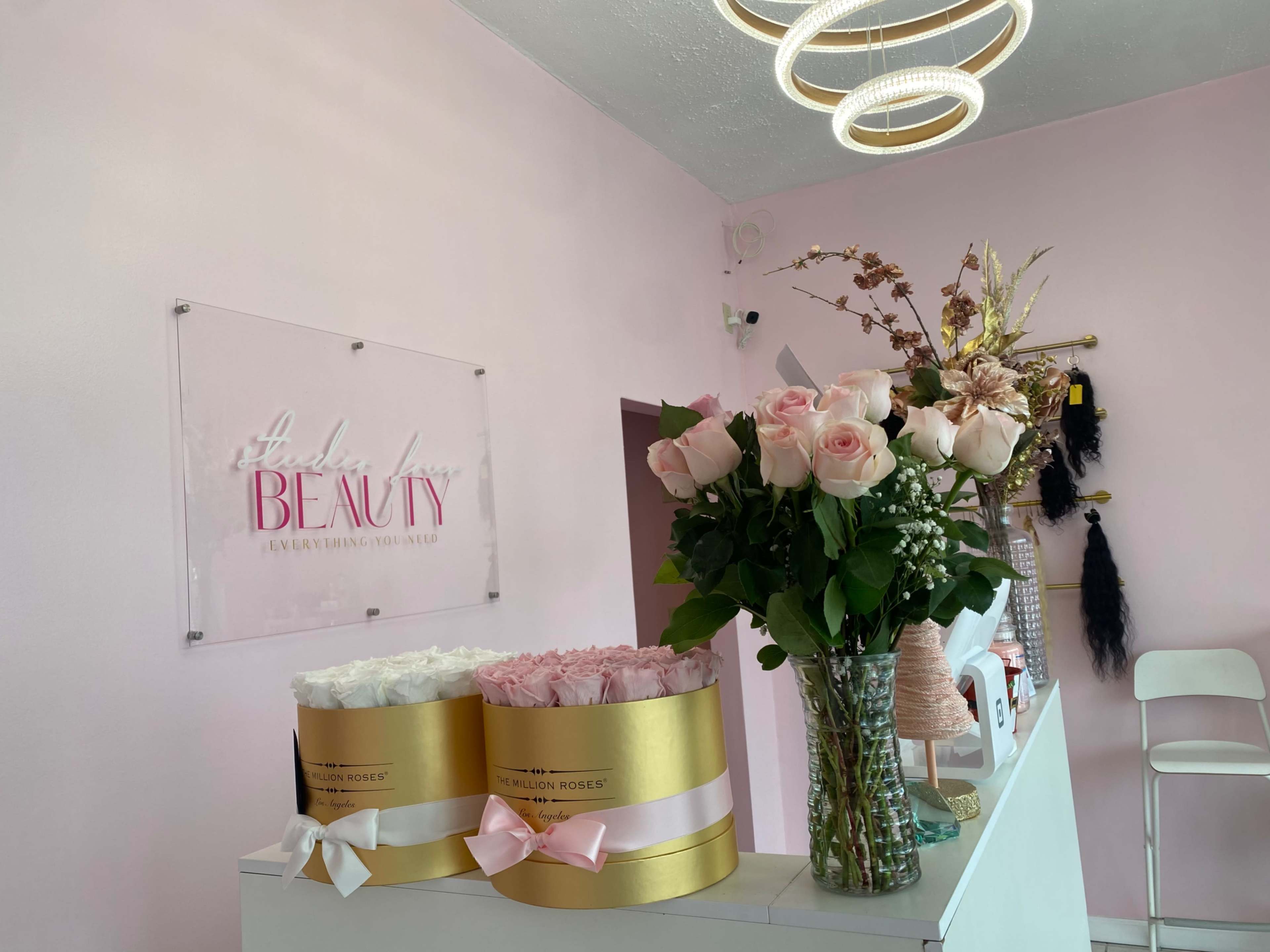 A beauty salon interior featuring a pink wall, a sign that reads "Salon of Beauty," and a counter adorned with two cylindrical flower arrangements and a vase of roses.