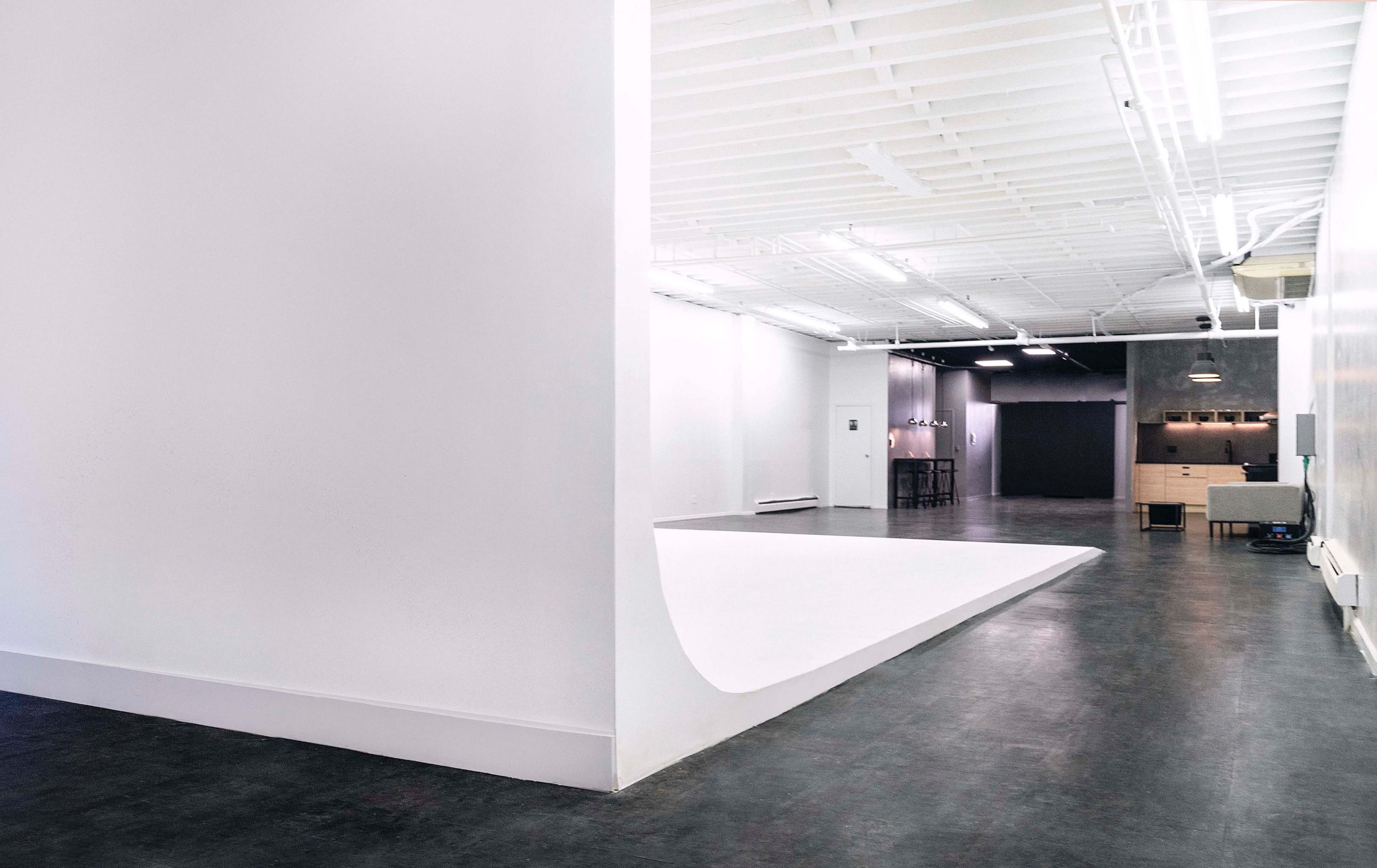 A spacious room with white walls, a curved white platform, and a dark area in the background featuring a kitchen setup.