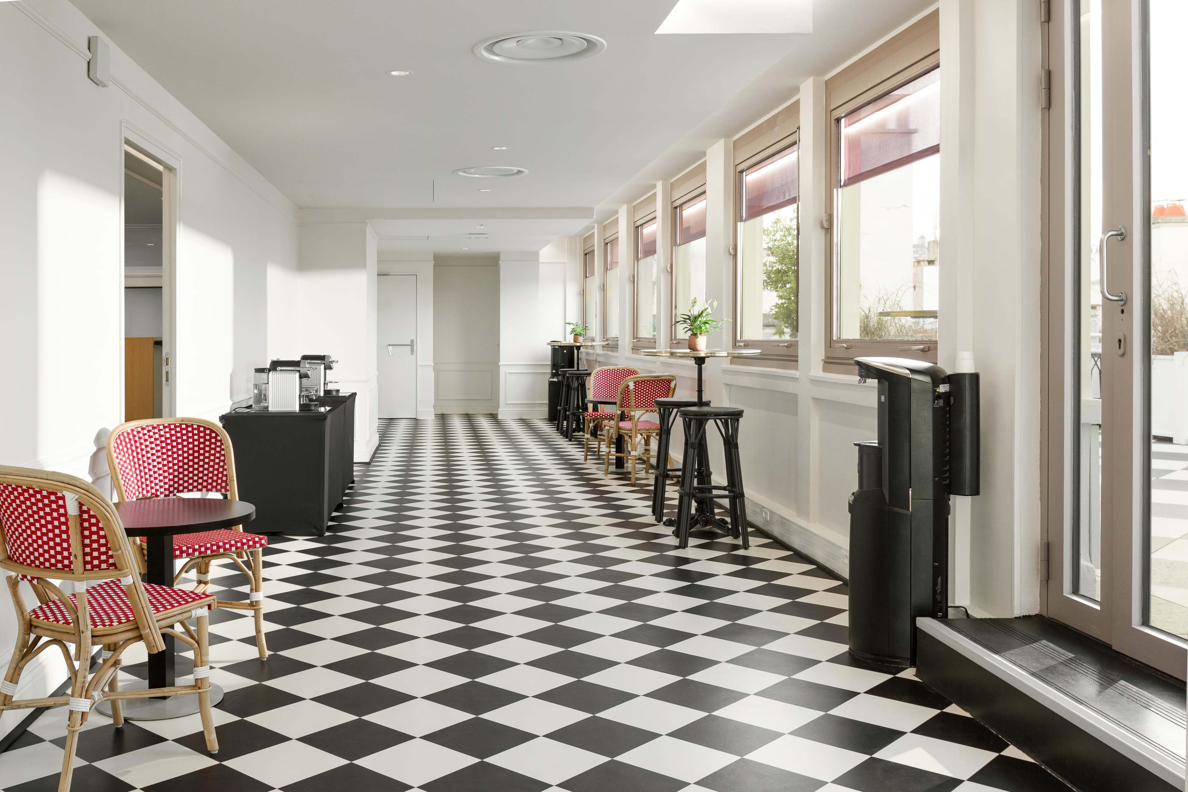The image shows a corridor with a black and white checkered floor, lined with windows, chairs, and a coffee station.