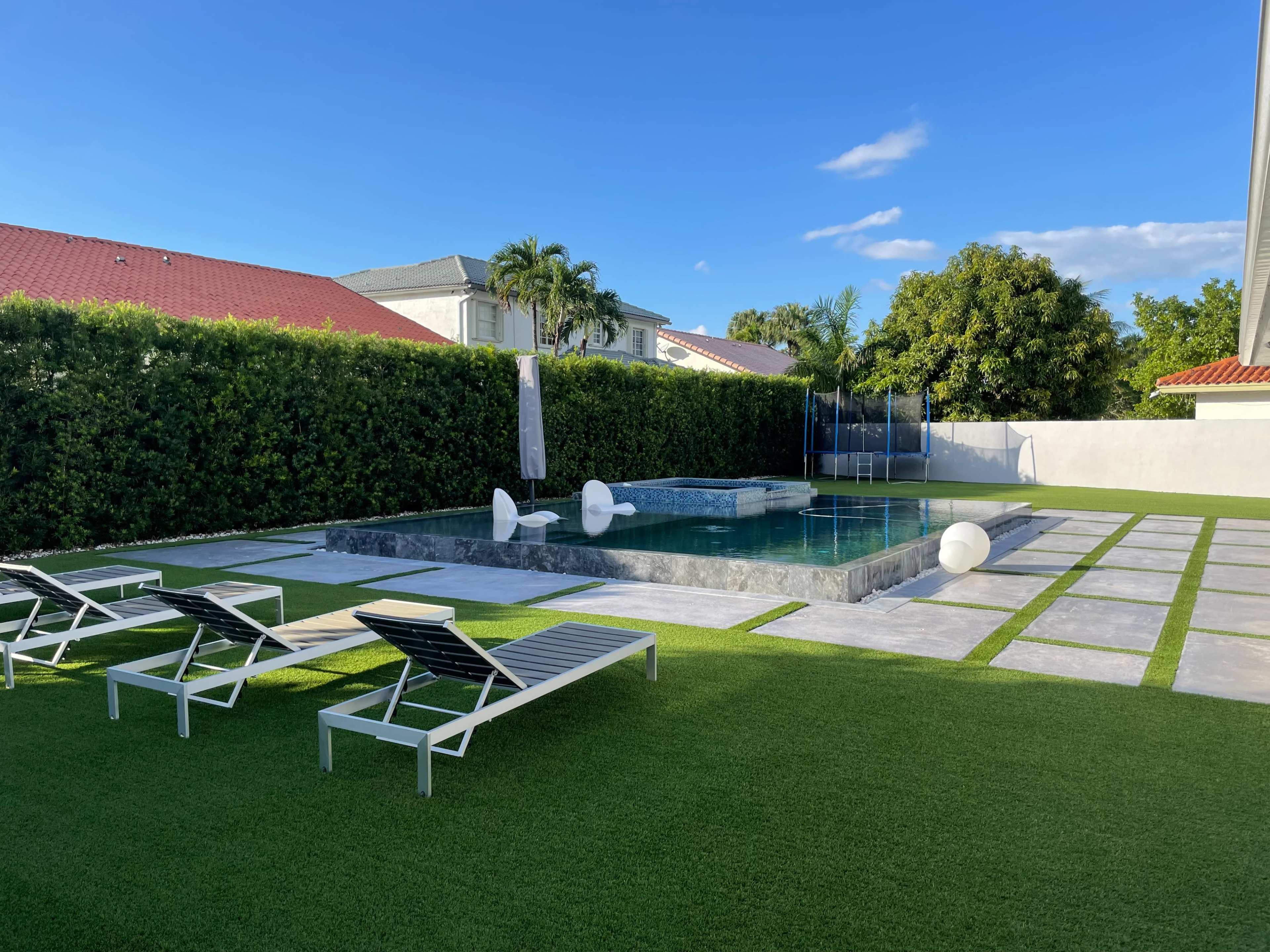 The image shows a modern backyard with a swimming pool surrounded by neatly arranged lounge chairs and a well-maintained lawn.