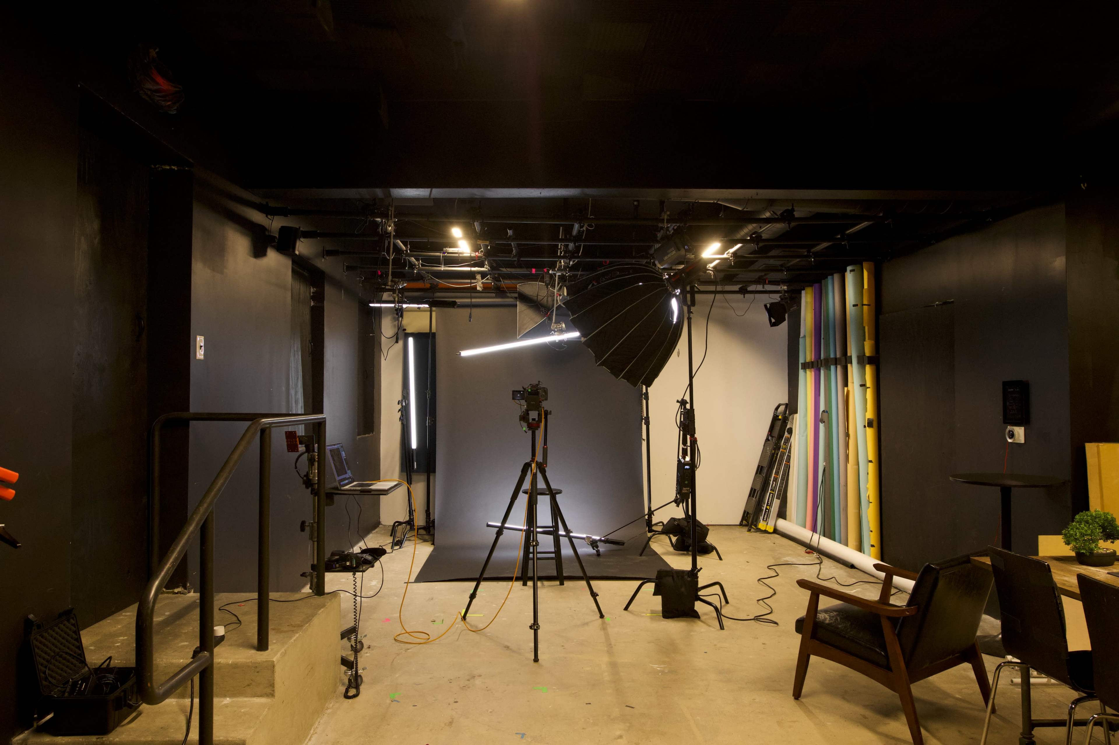 The image shows a photography studio featuring a camera on a tripod, a large softbox light, and various equipment against a backdrop with colorful vertical stripes.