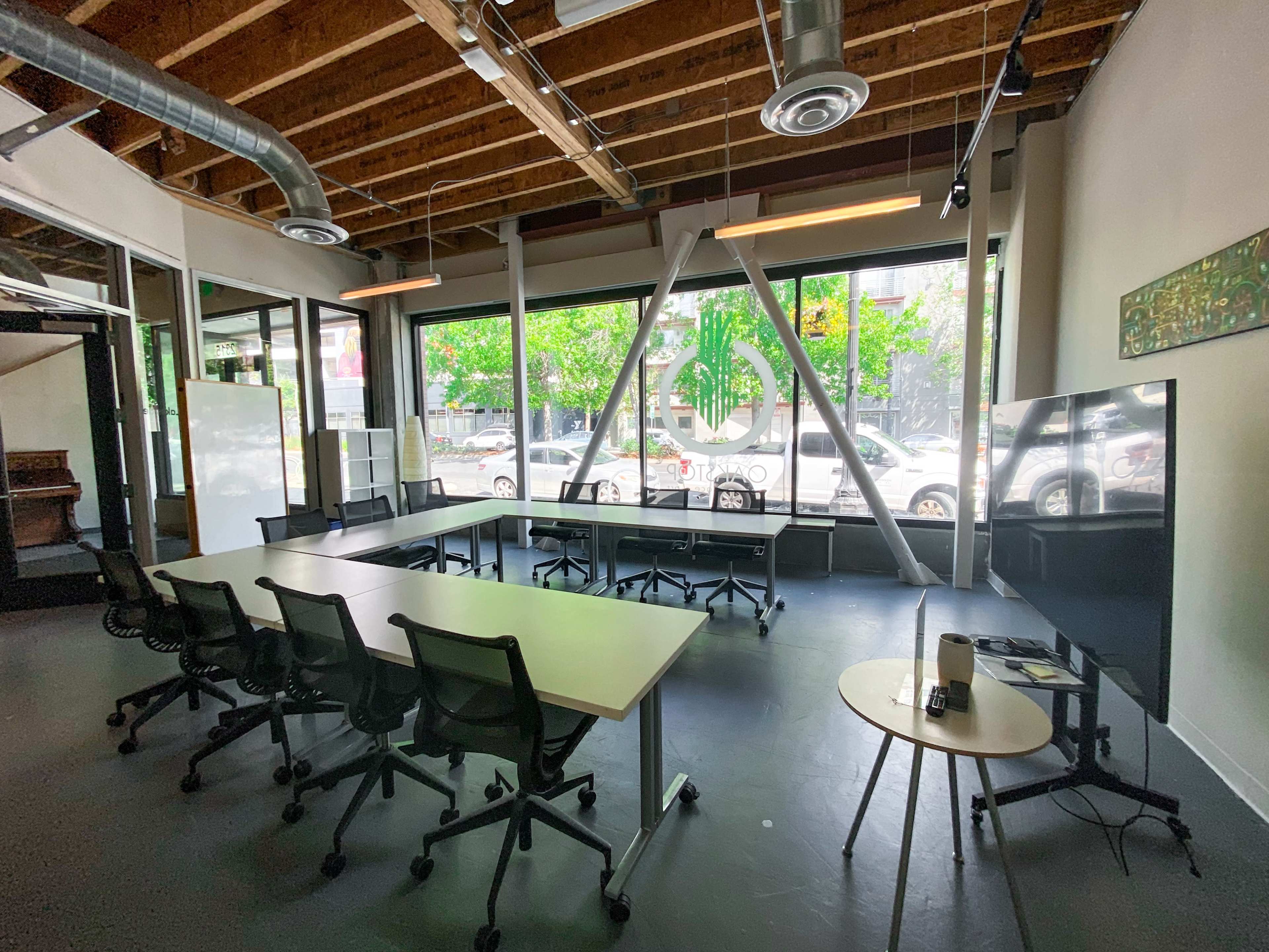 A modern office space features a long table surrounded by six chairs, large windows with greenery outside, and a television mounted on the wall.