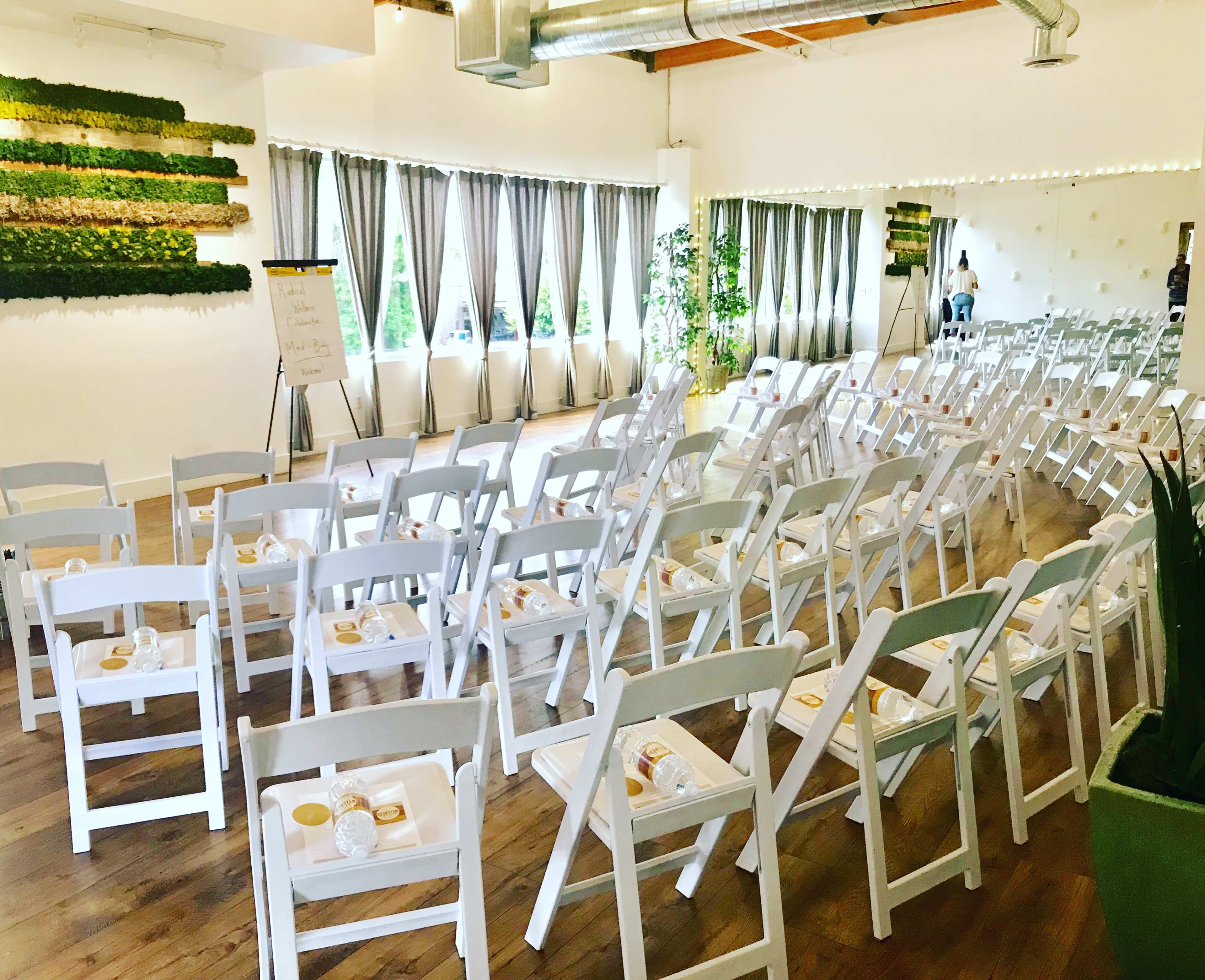A spacious event room arranged with rows of white chairs, each with a notepad and snack on the seat, and large windows draped with gray curtains.