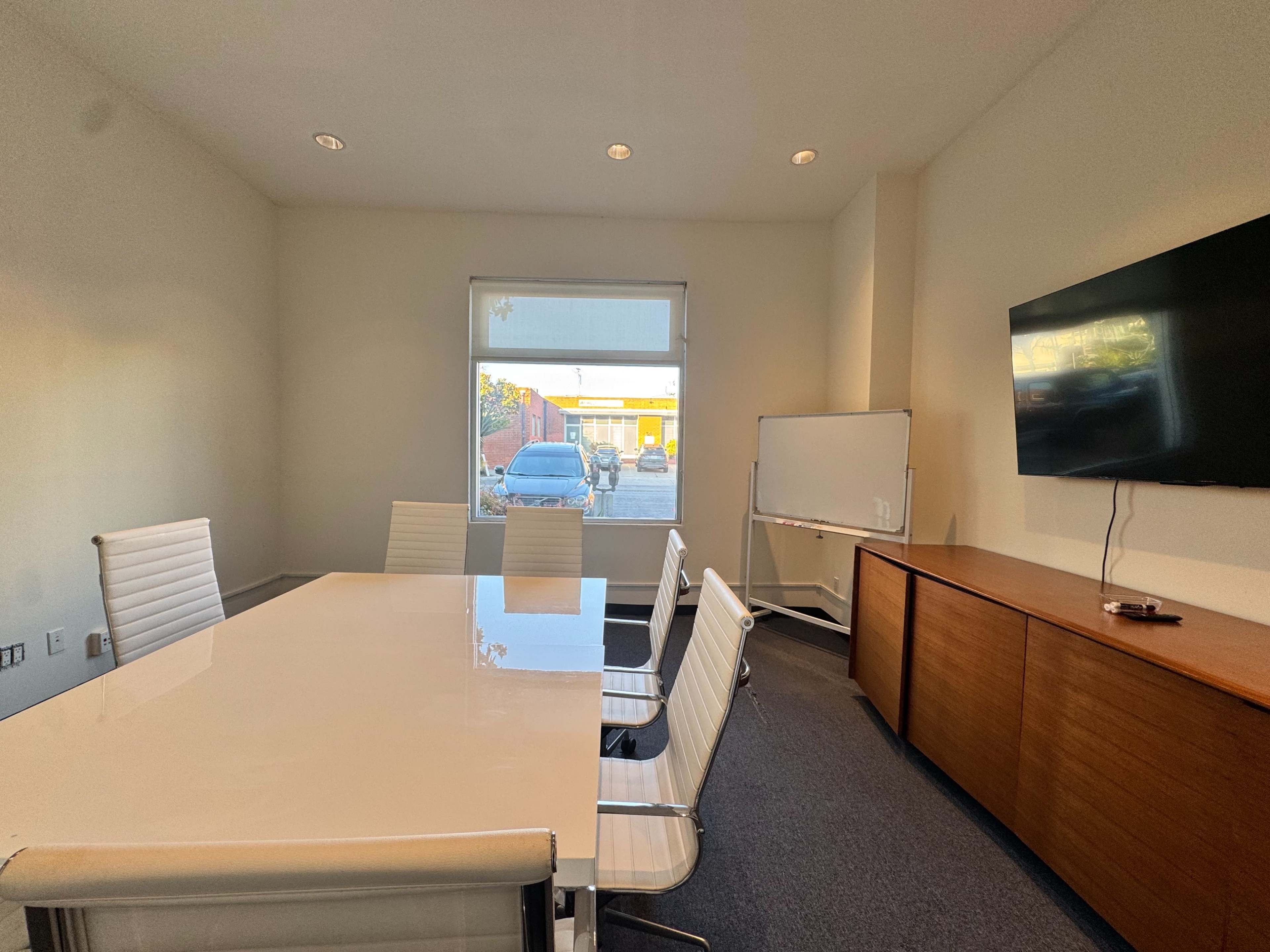 The image shows a modern conference room with a large white table surrounded by several white chairs, a television mounted on the wall, and a window providing a view of the outside.