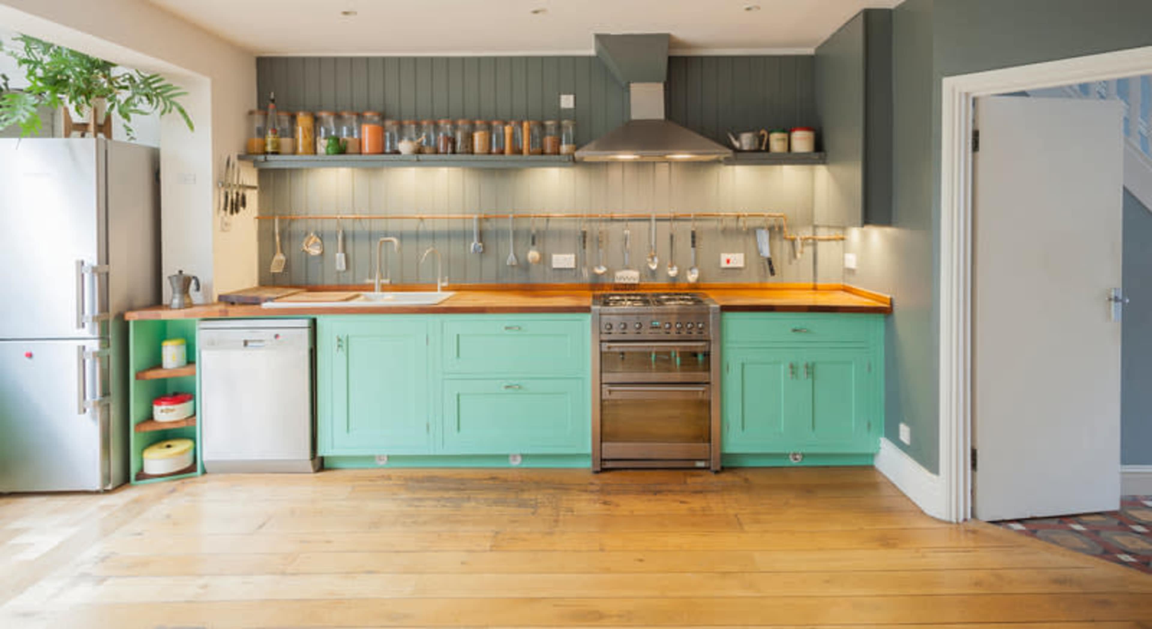 A kitchen features mint green cabinetry, a stainless steel stove, and a wooden countertop, with metal shelves displaying various kitchenware.