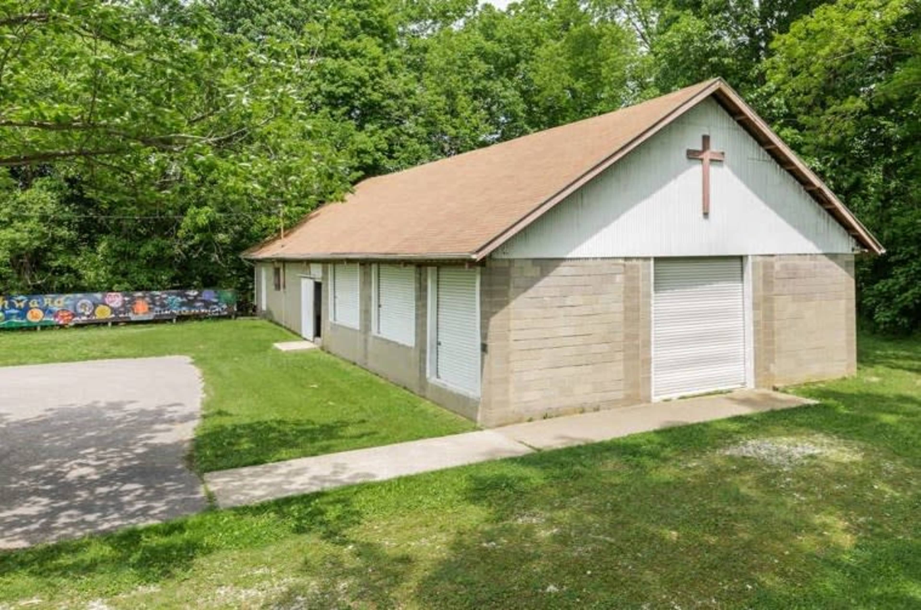 A single-story building with a brown roof, a white cross on the front, and multiple shuttered windows, surrounded by grass and trees.