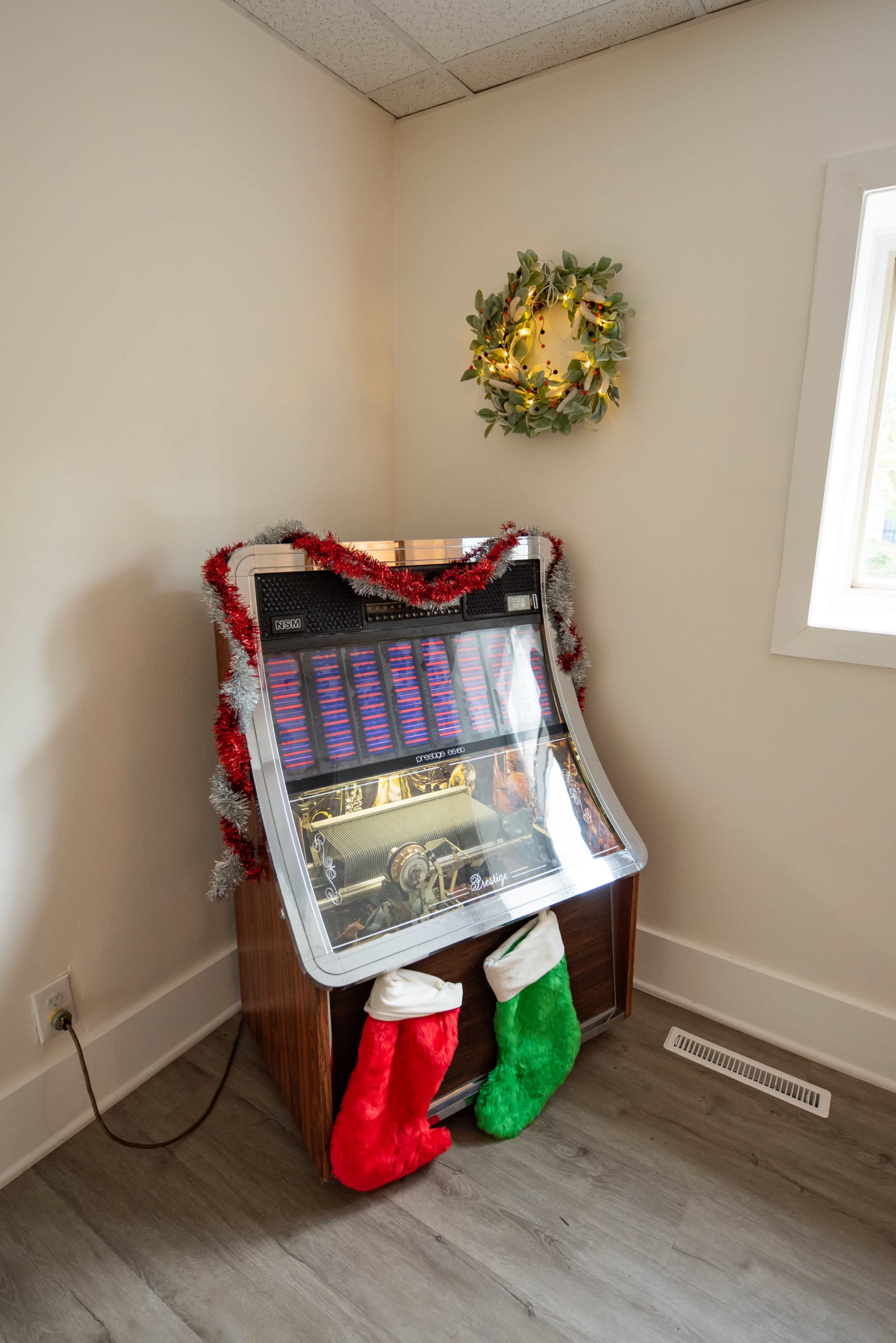 A jukebox is adorned with festive decorations, including tinsel and holiday stockings, in a corner of a brightly lit room.