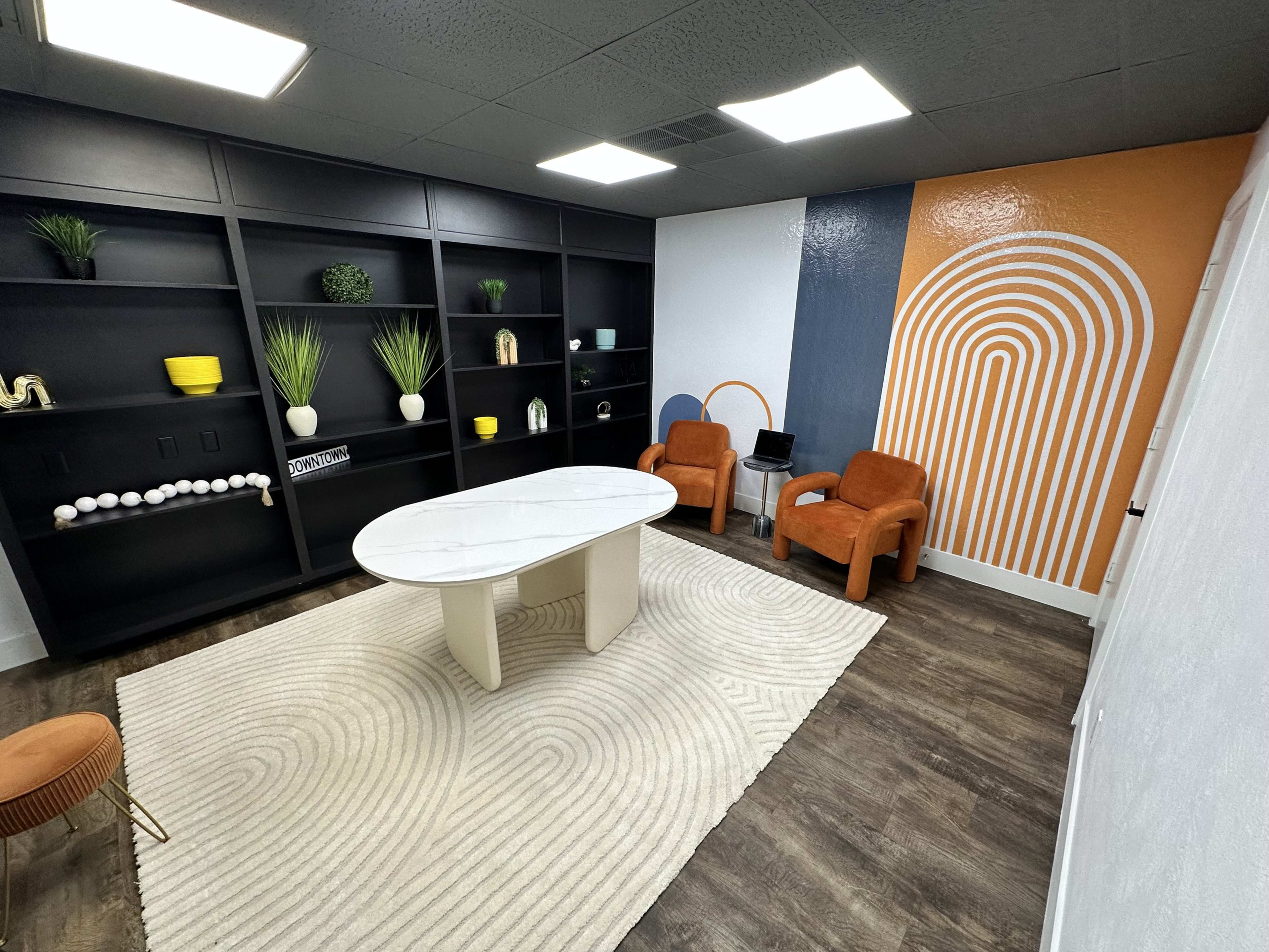 The image shows a modern office space featuring a round marble table, two orange chairs, a black bookshelf with decorative items, and a colorful wall with abstract designs.