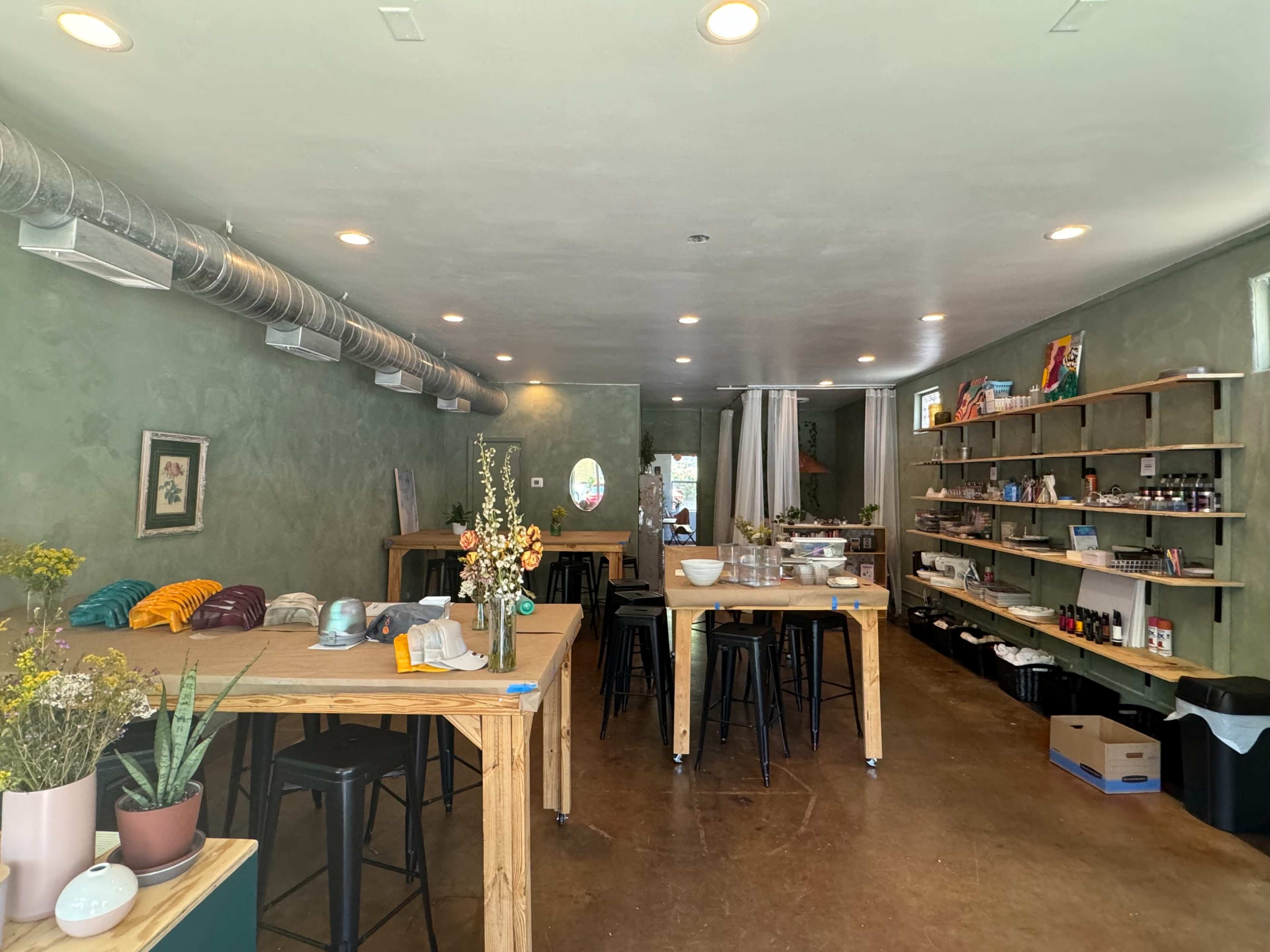 The image shows a spacious interior of a shop featuring wooden tables, shelves stocked with various items, and green walls.