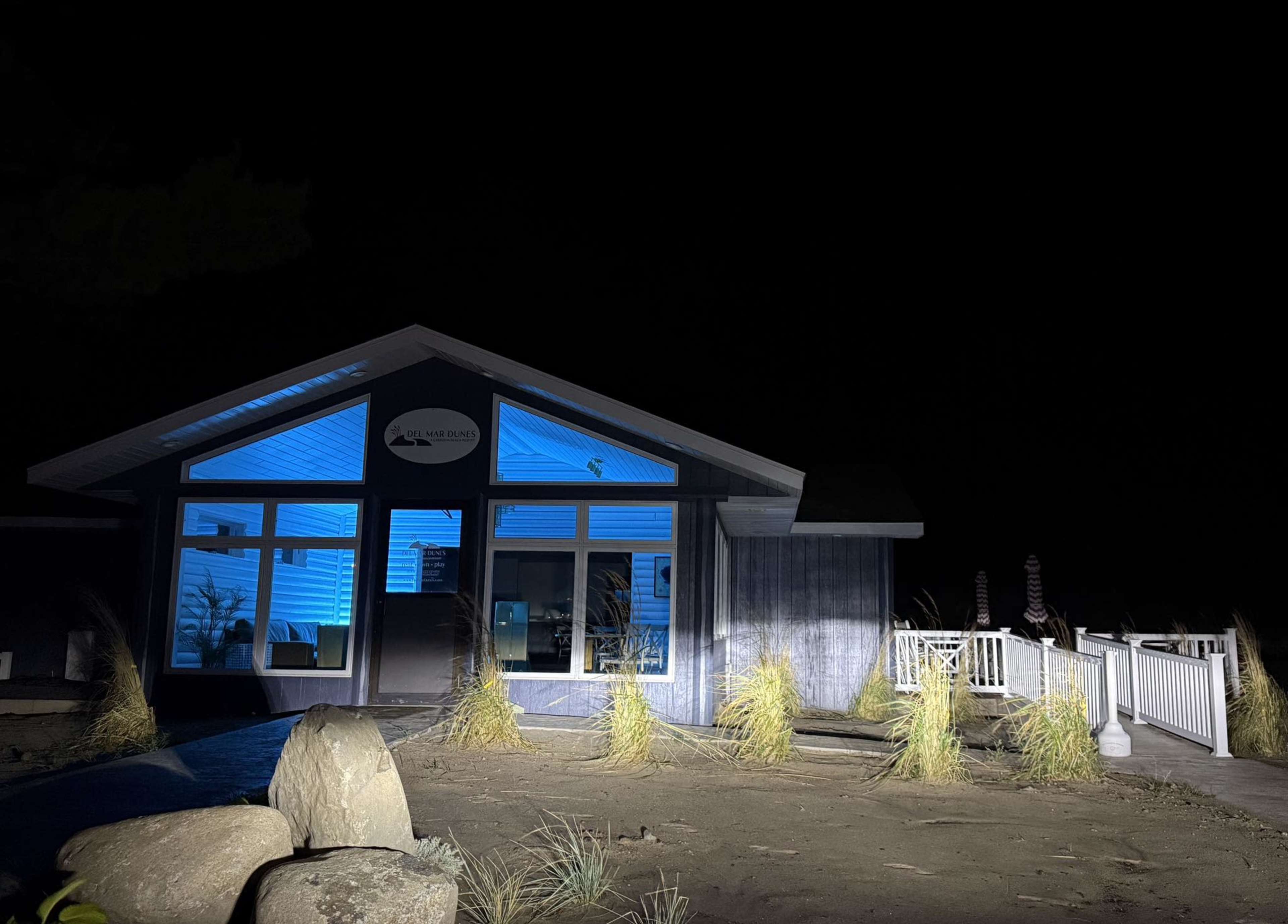 A coastal house is illuminated by blue lights at night, surrounded by sandy landscaping and rocks.