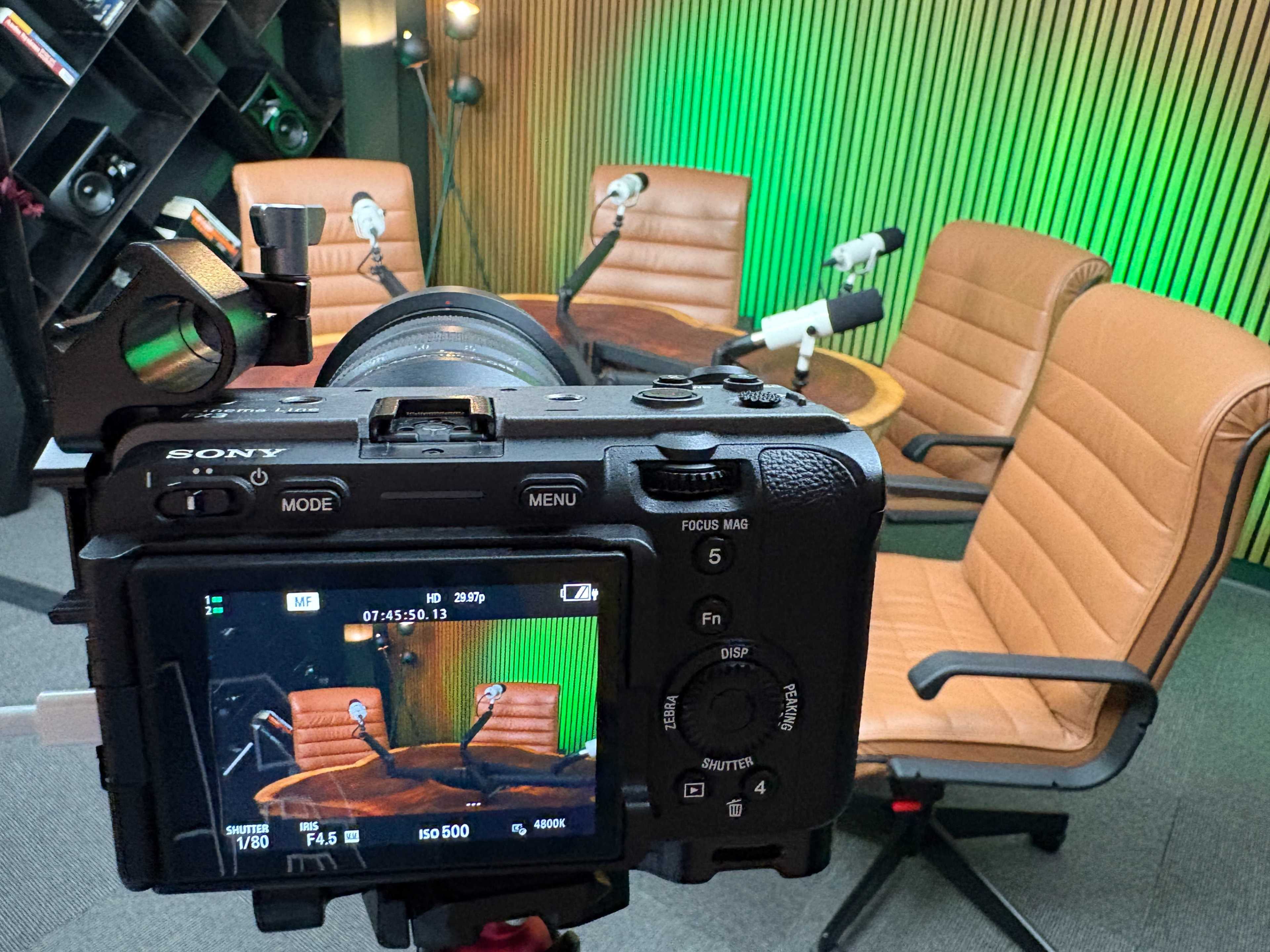 The image shows a camera focused on a well-lit podcast recording setup with two brown chairs, microphones, and a wooden table against a green-lit background.