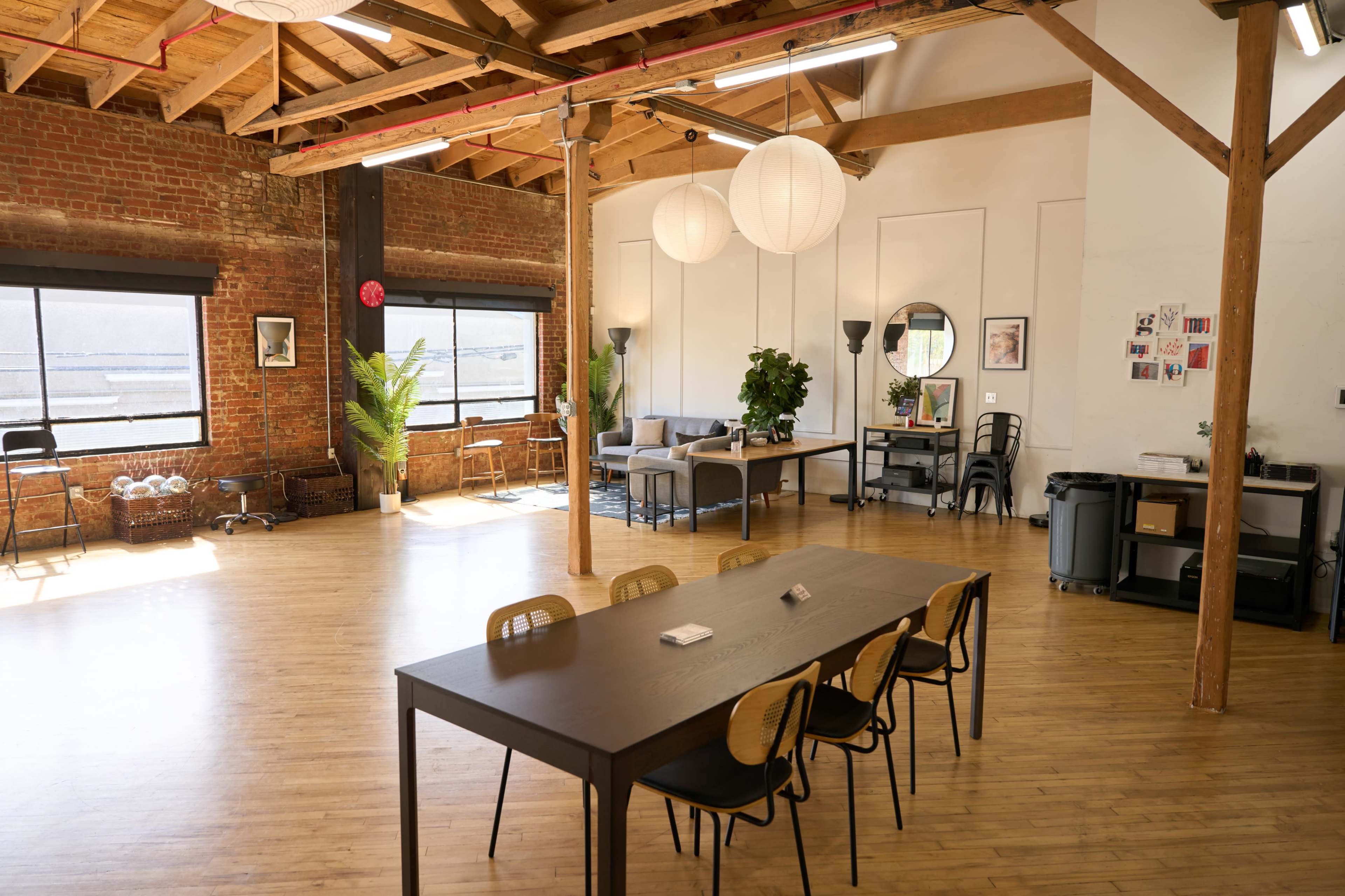 The image shows a spacious, well-lit indoor area with wooden flooring, large windows, a long table surrounded by chairs, and various plants and seating arrangements.