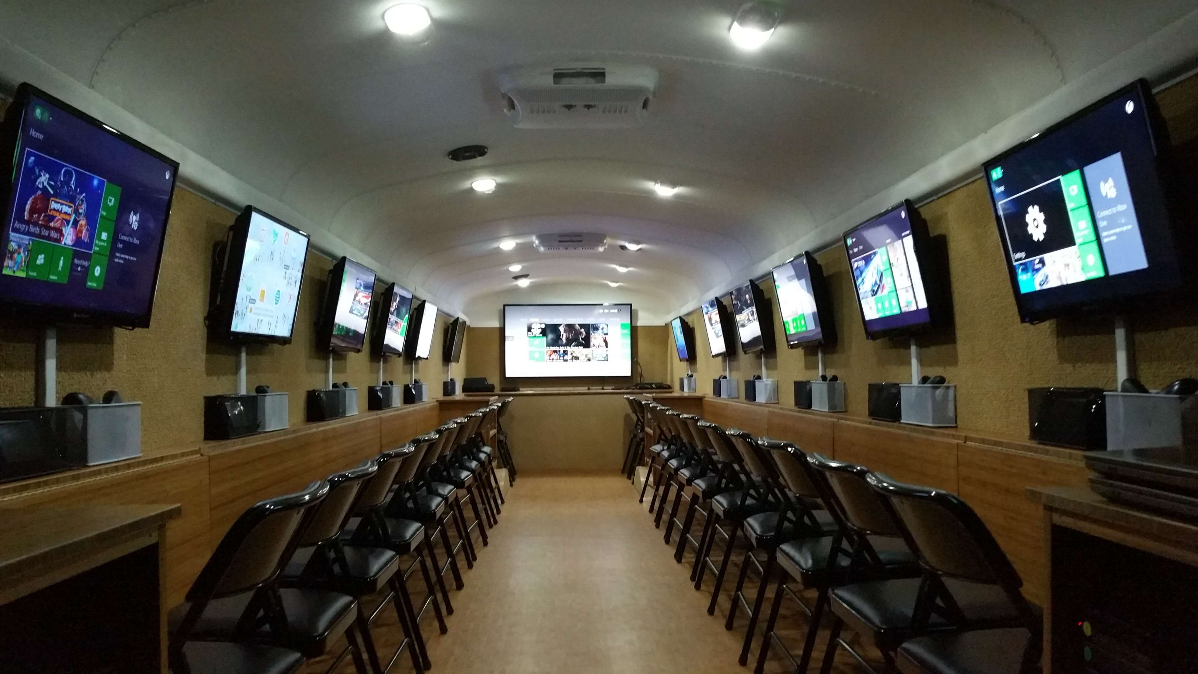 A mobile classroom is arranged with multiple screens along the walls and rows of chairs facing a central display.
