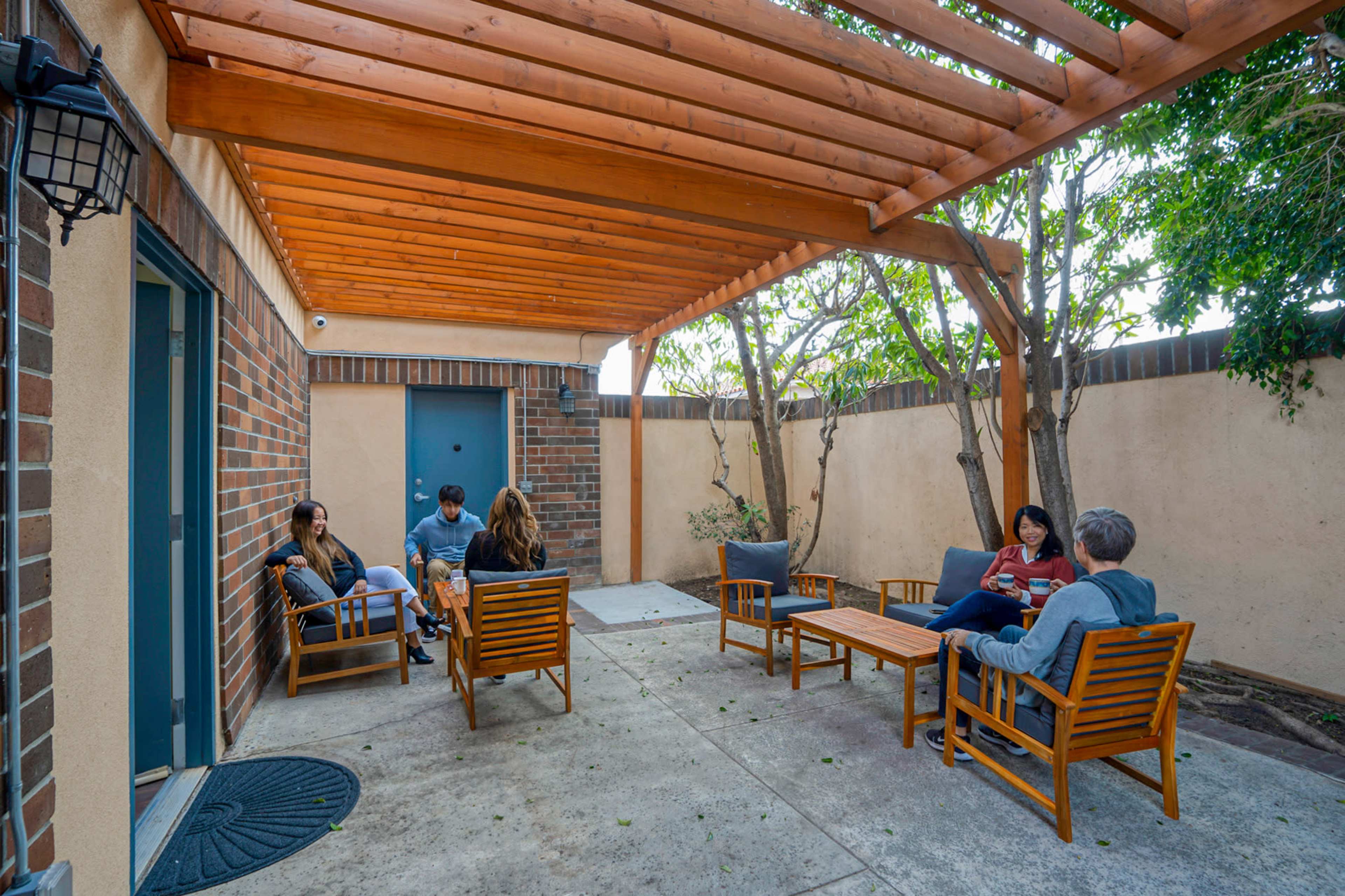 A group of five people is seated on wooden furniture under a wooden pergola in a small outdoor patio area.