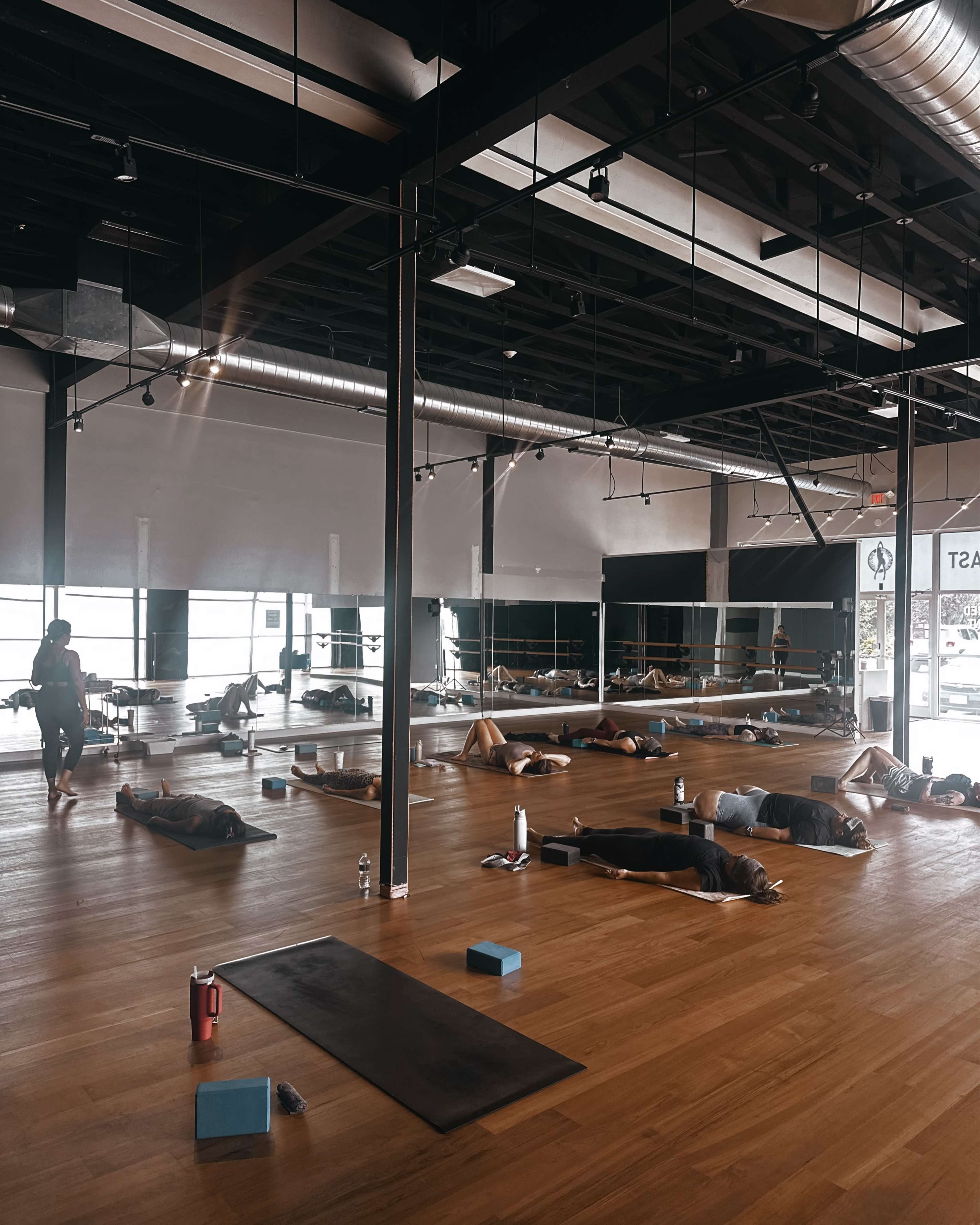A spacious fitness studio features several individuals practicing yoga on mats, with large windows providing natural light.