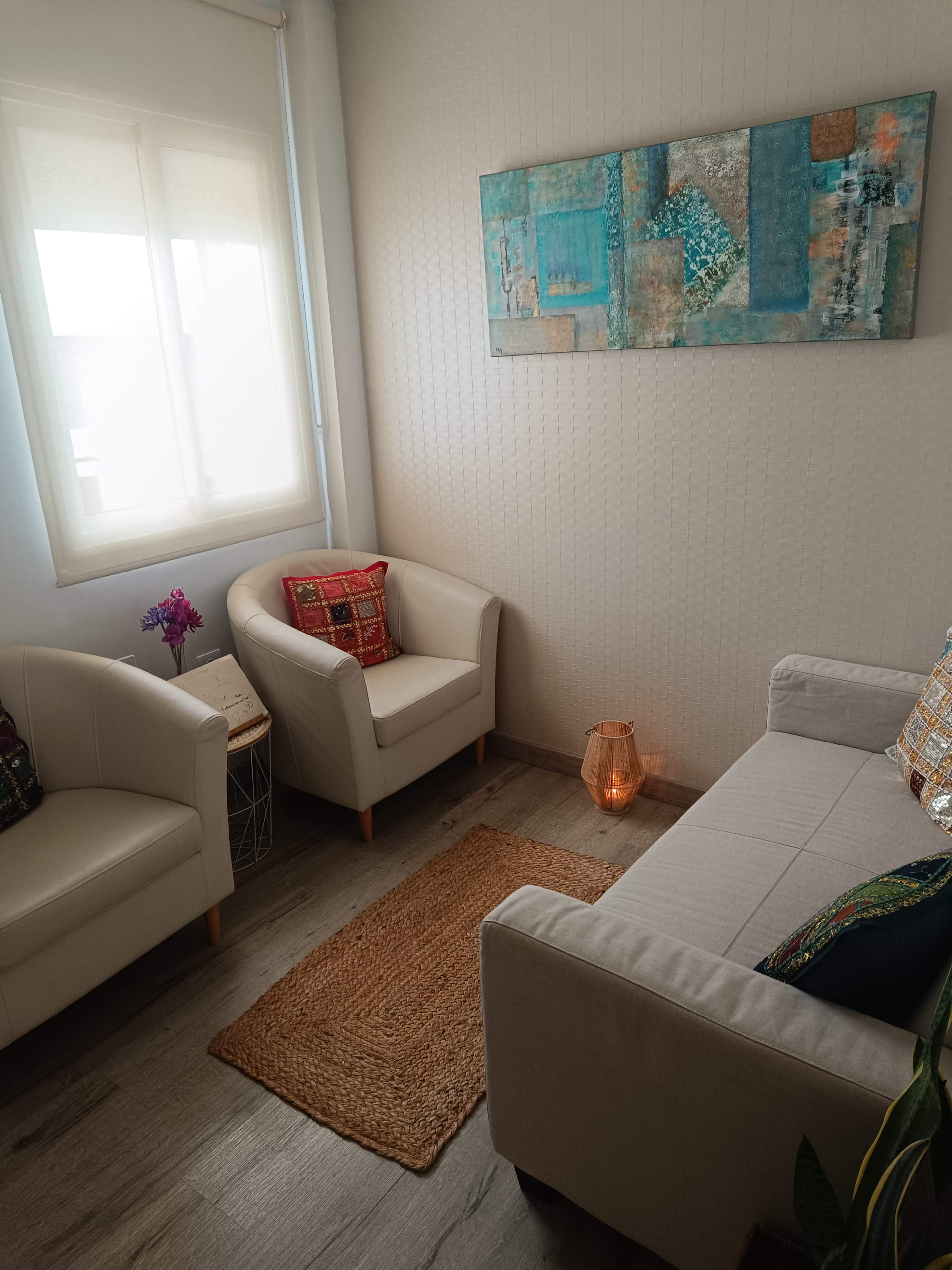 The image shows a cozy living area with two white armchairs, a light-colored sofa, a woven rug, and a colorful wall hanging above the sofa.