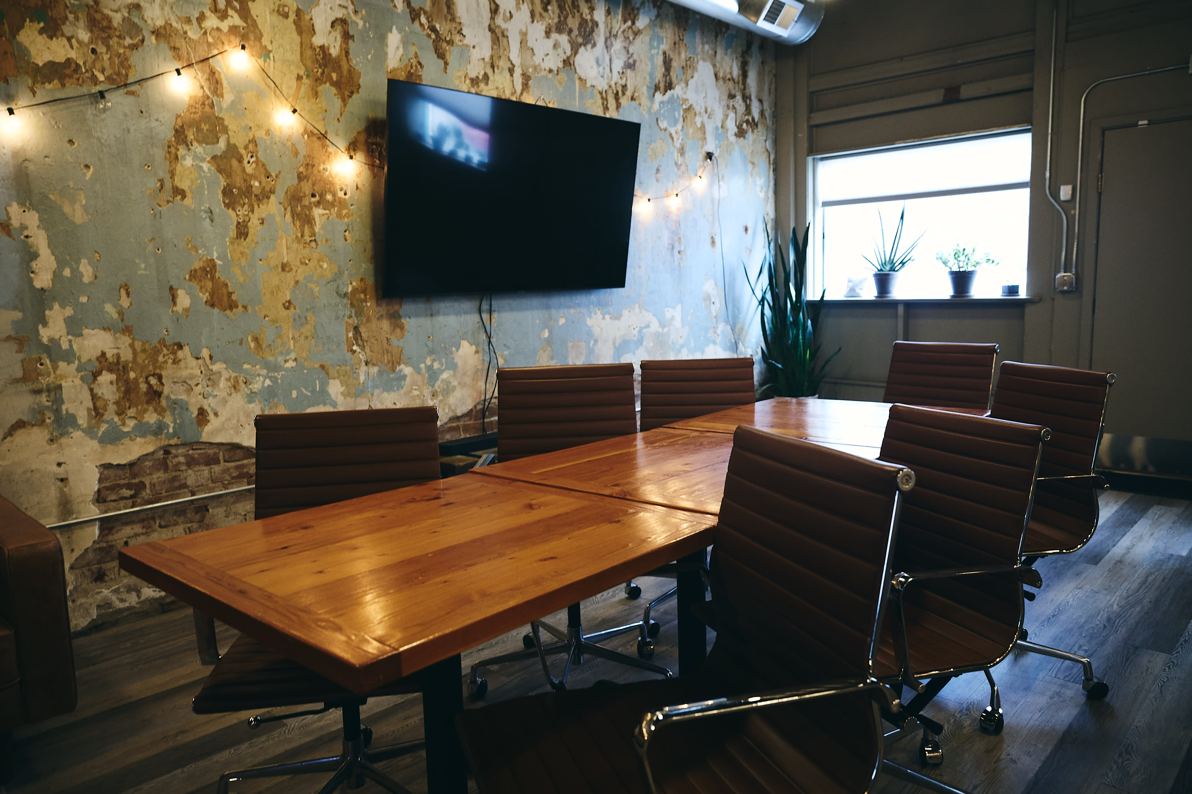 A conference room features a long wooden table surrounded by brown leather chairs, with a large TV mounted on a weathered blue wall and plants visible in the window.