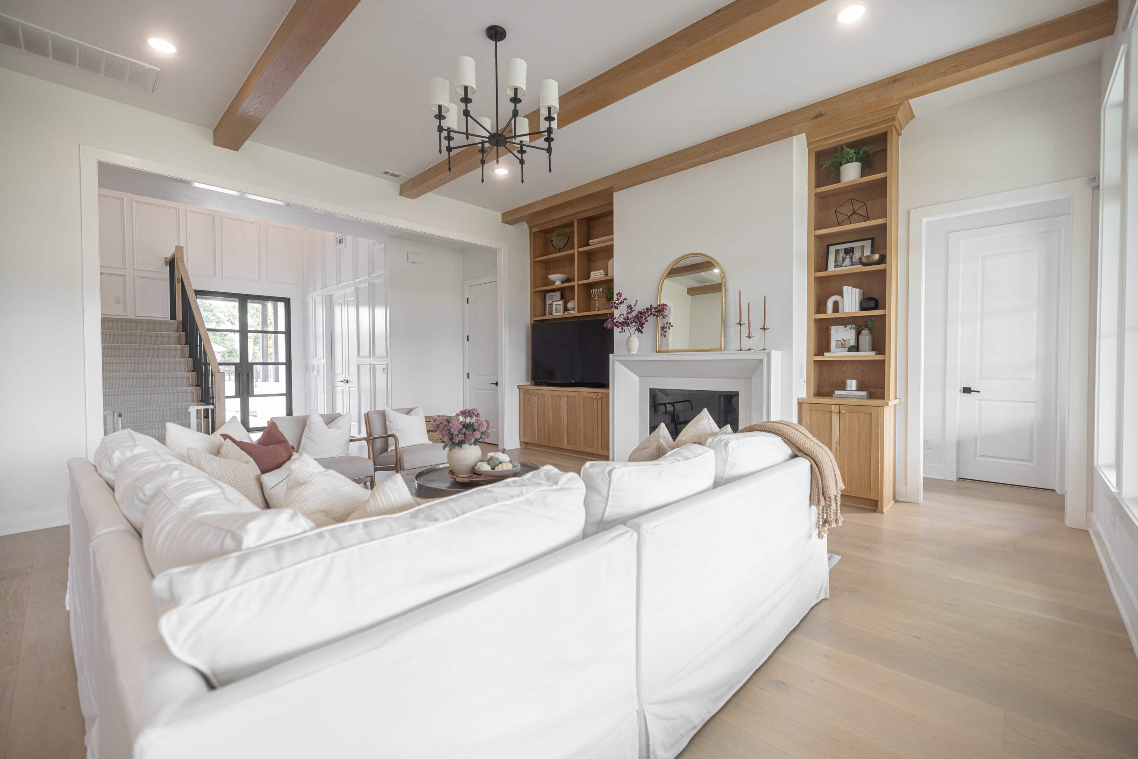 A spacious living room featuring a white sectional sofa, wooden beams across the ceiling, and a fireplace with built-in shelves on either side.
