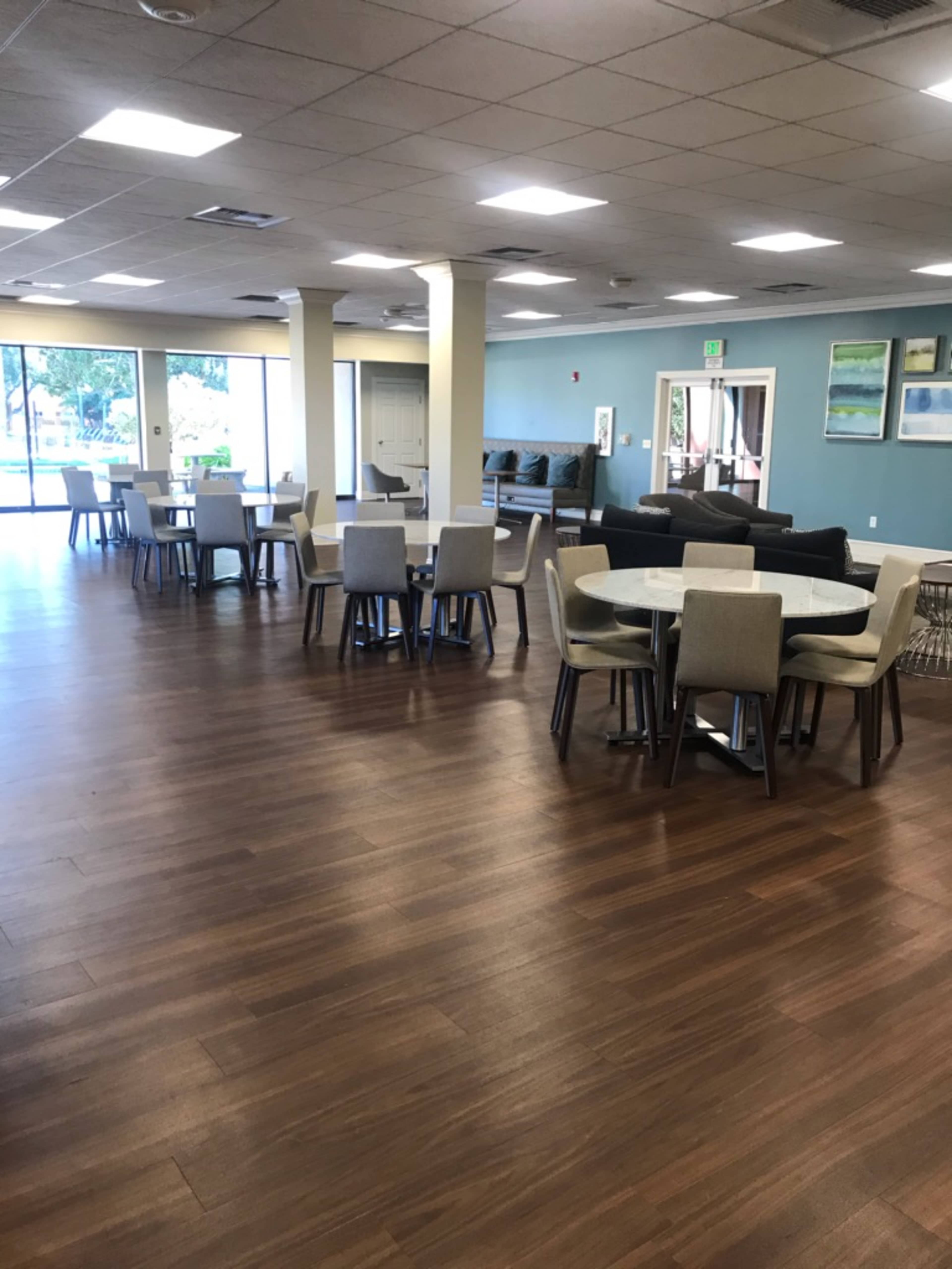 A spacious communal area with several round and rectangular tables surrounded by chairs, and a lounge area in the background.