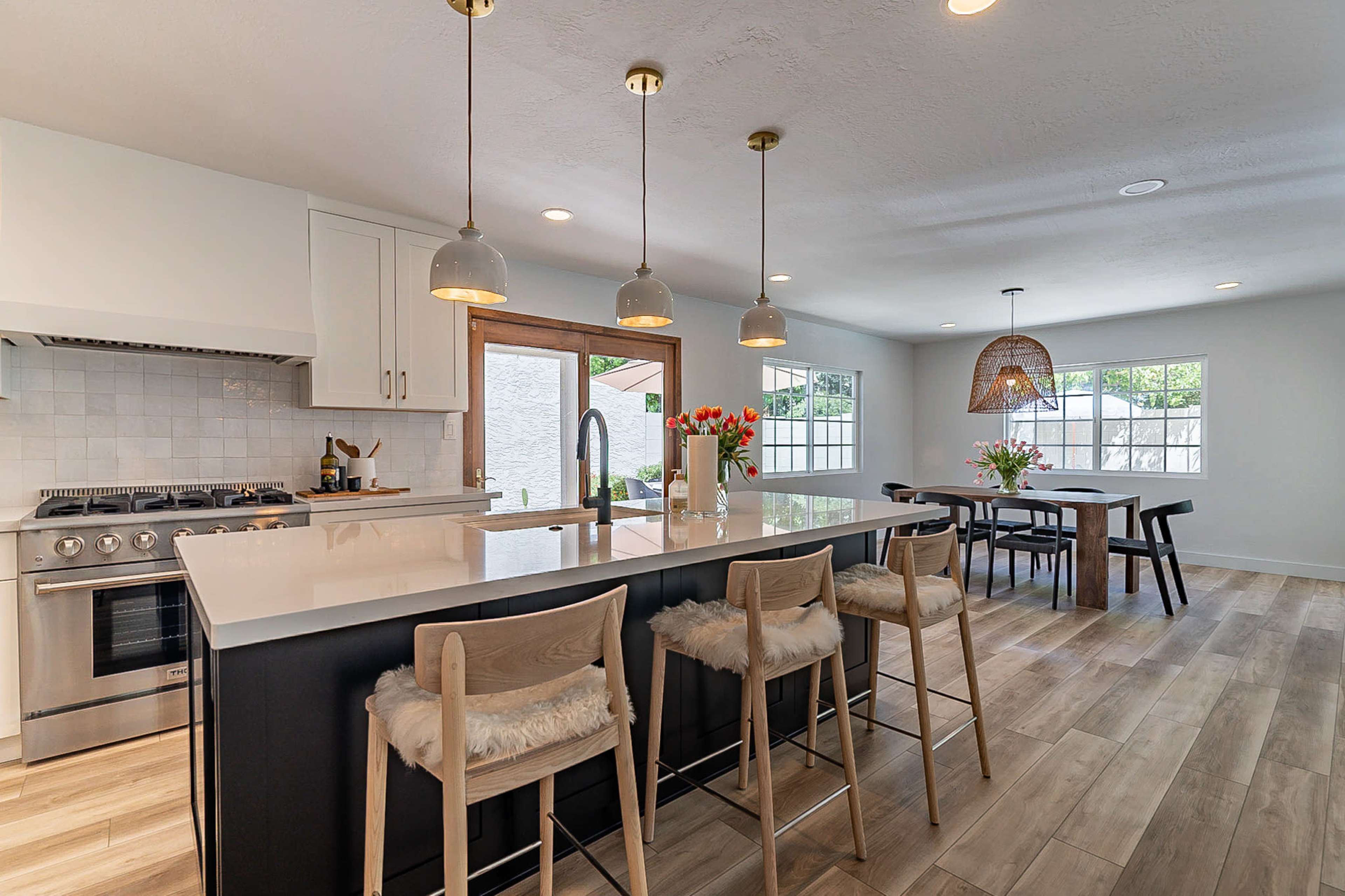 A modern kitchen features a large island with seating, pendant lights, and a dining area with a table and flower arrangements.