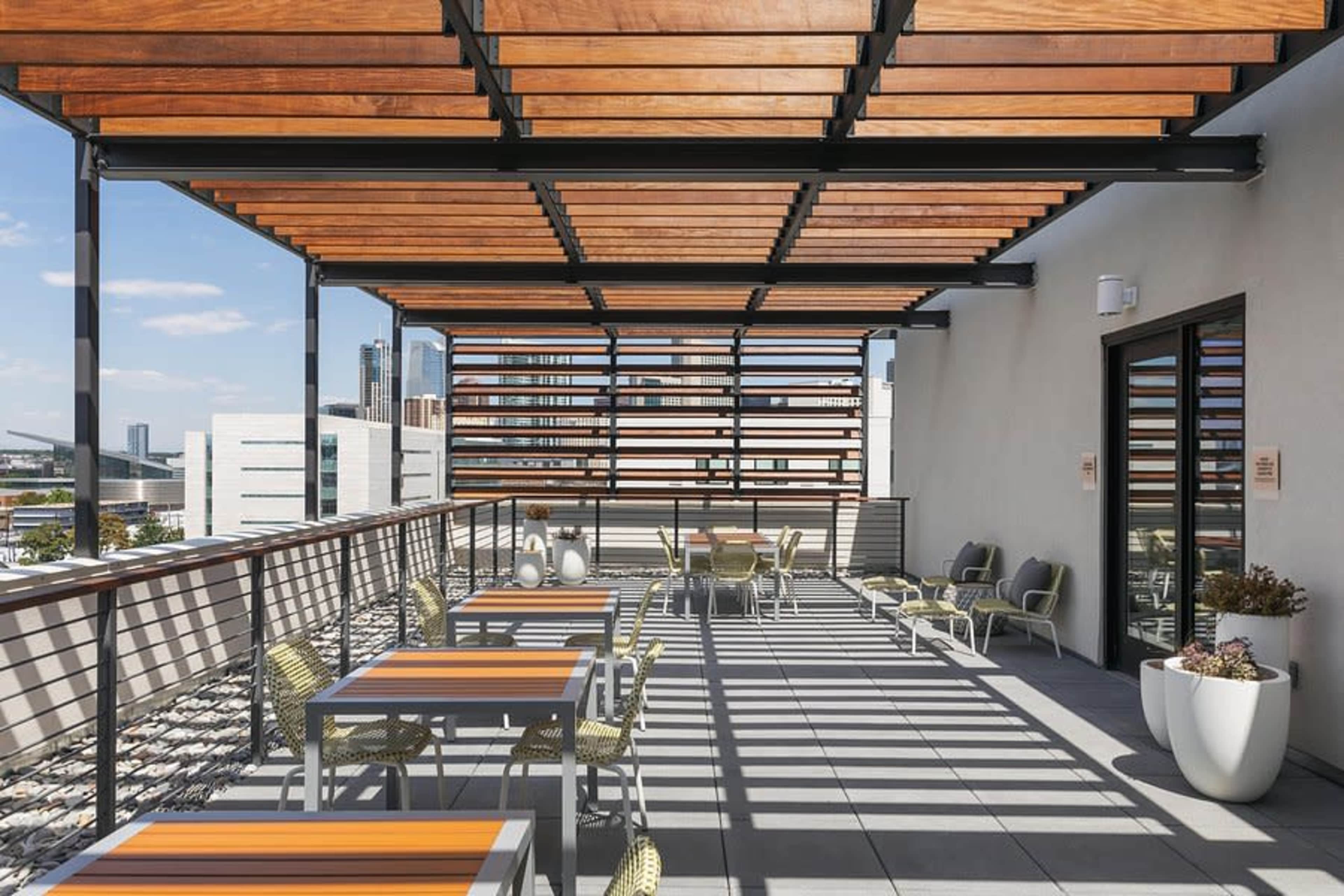 The image shows a modern rooftop terrace featuring wooden slatted awnings, tables with orange accents, and a view of city buildings in the background.