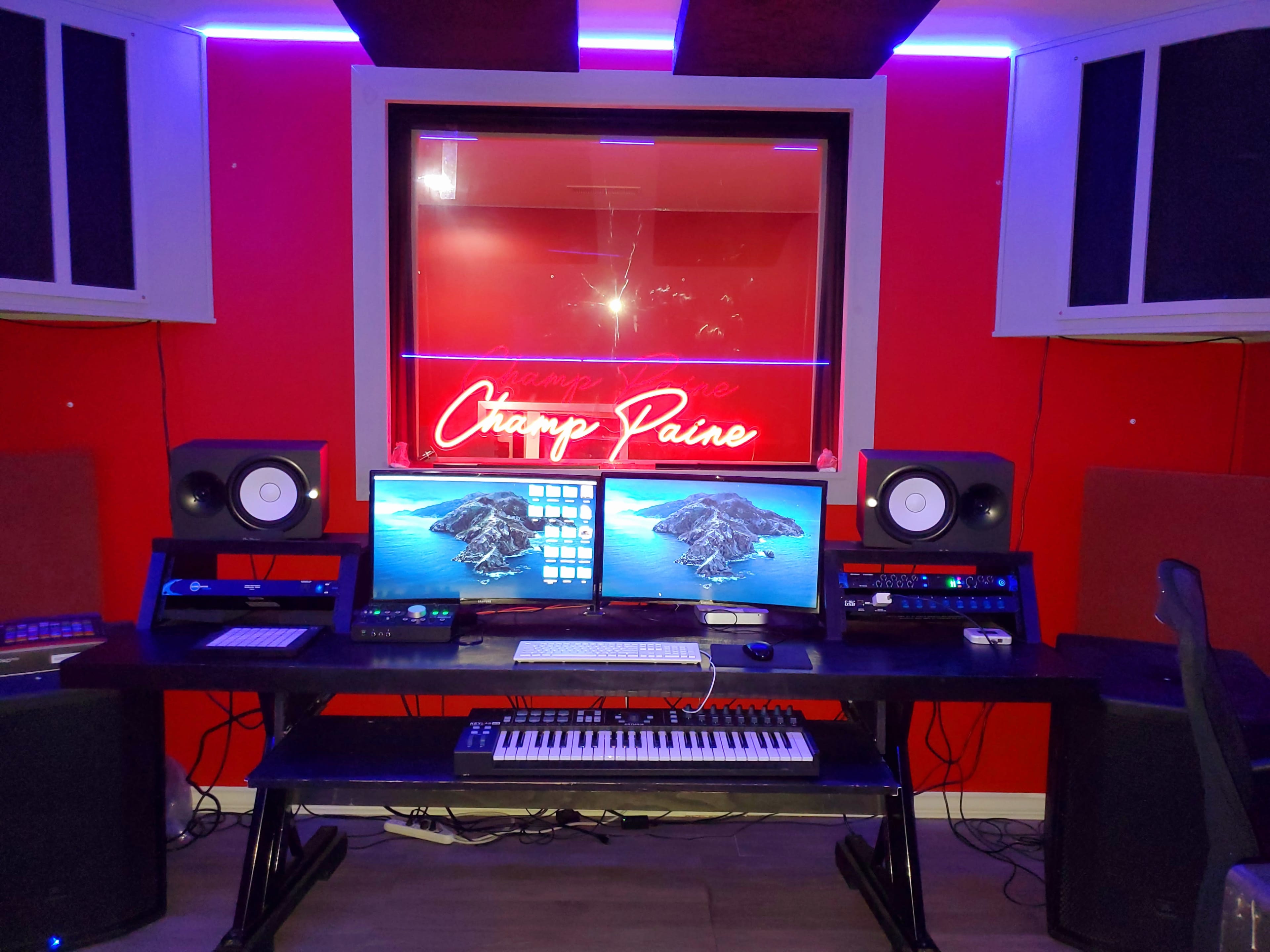 The image shows a music studio with two computers, a keyboard, and speakers, featuring red walls and illuminated signage.