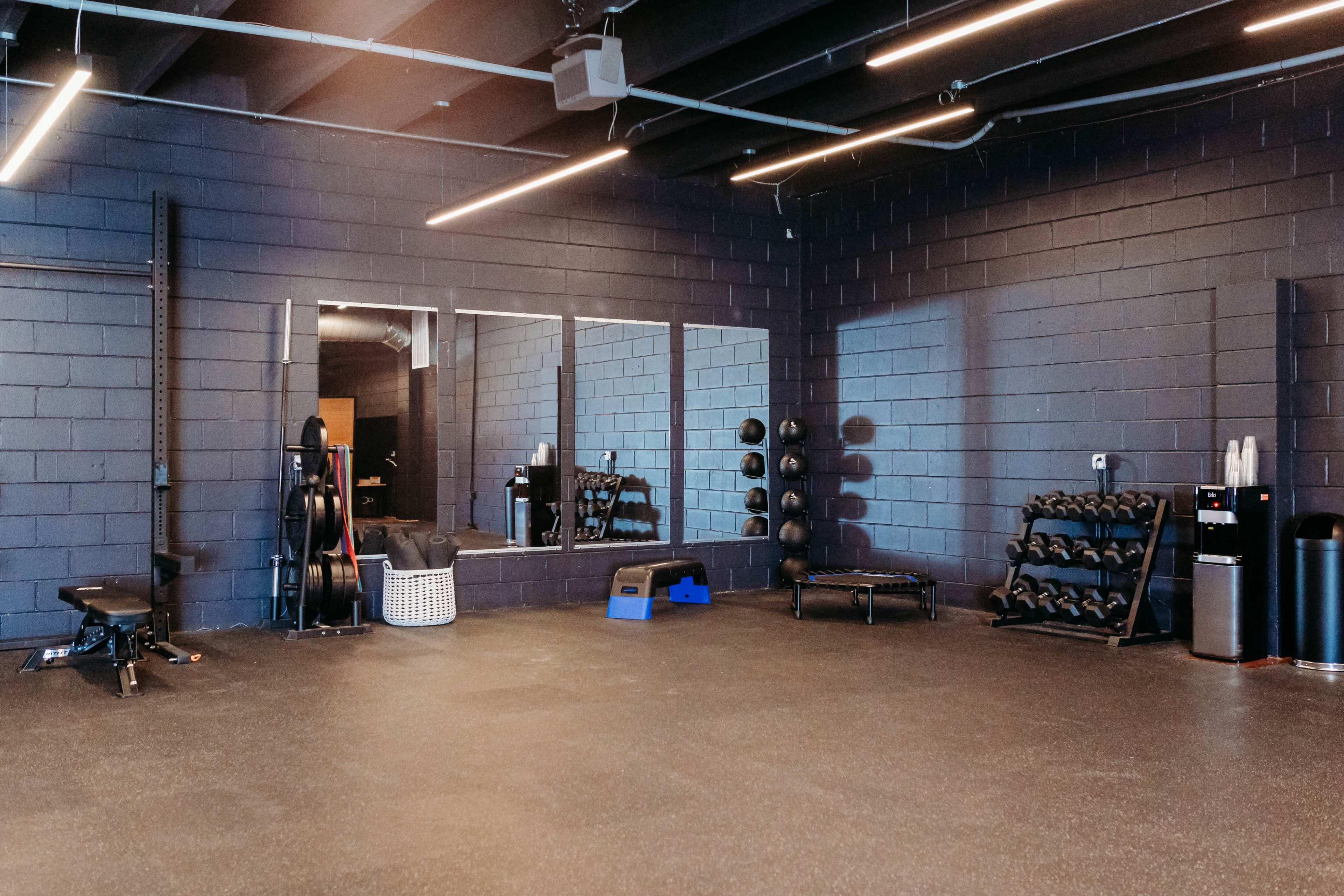 The image shows a spacious gym area featuring exercise equipment, mirrors, and free weights arranged against a black wall.