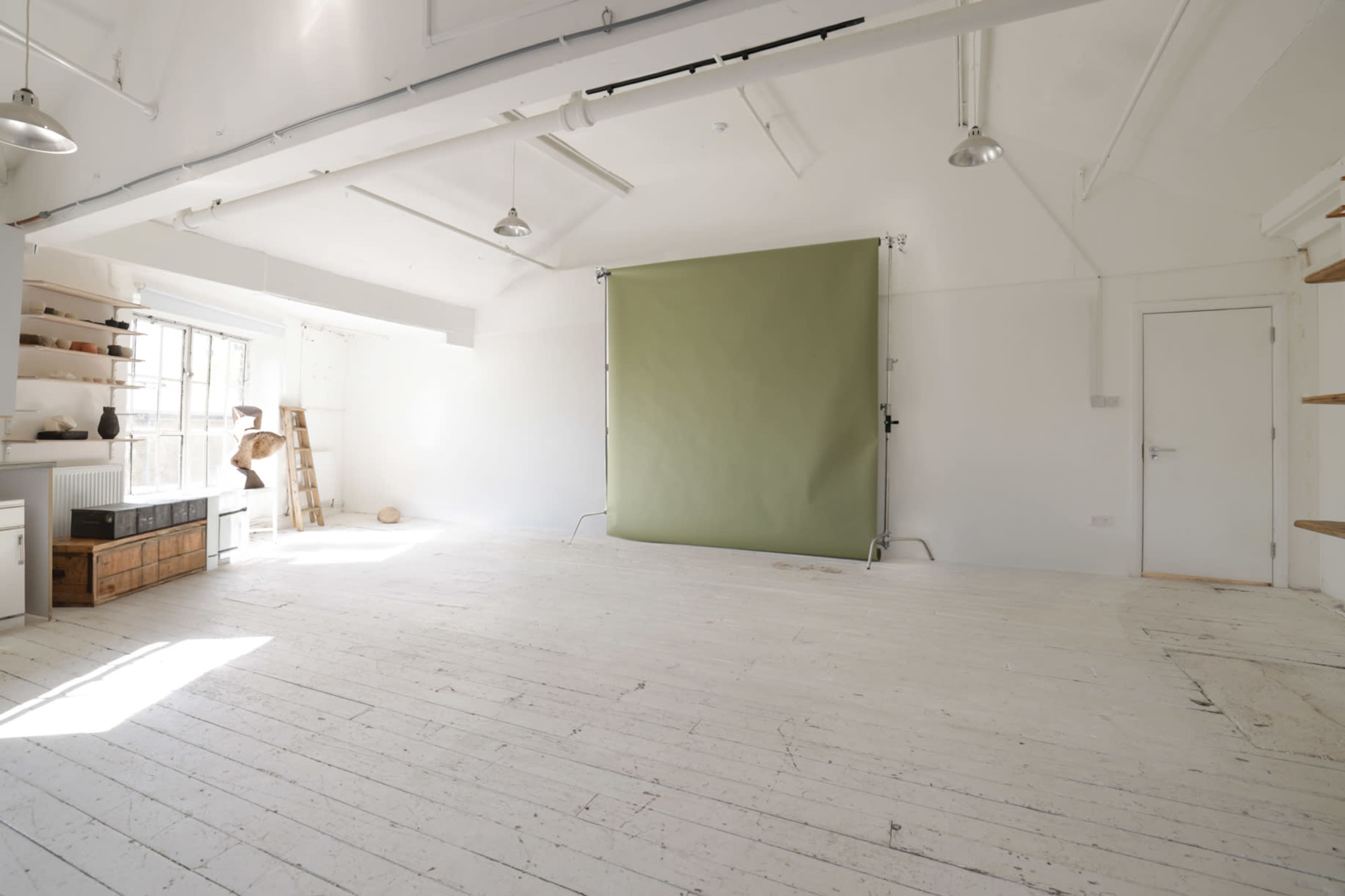 The image shows an empty, well-lit studio with white walls and a green backdrop, featuring wooden shelves and a large window.