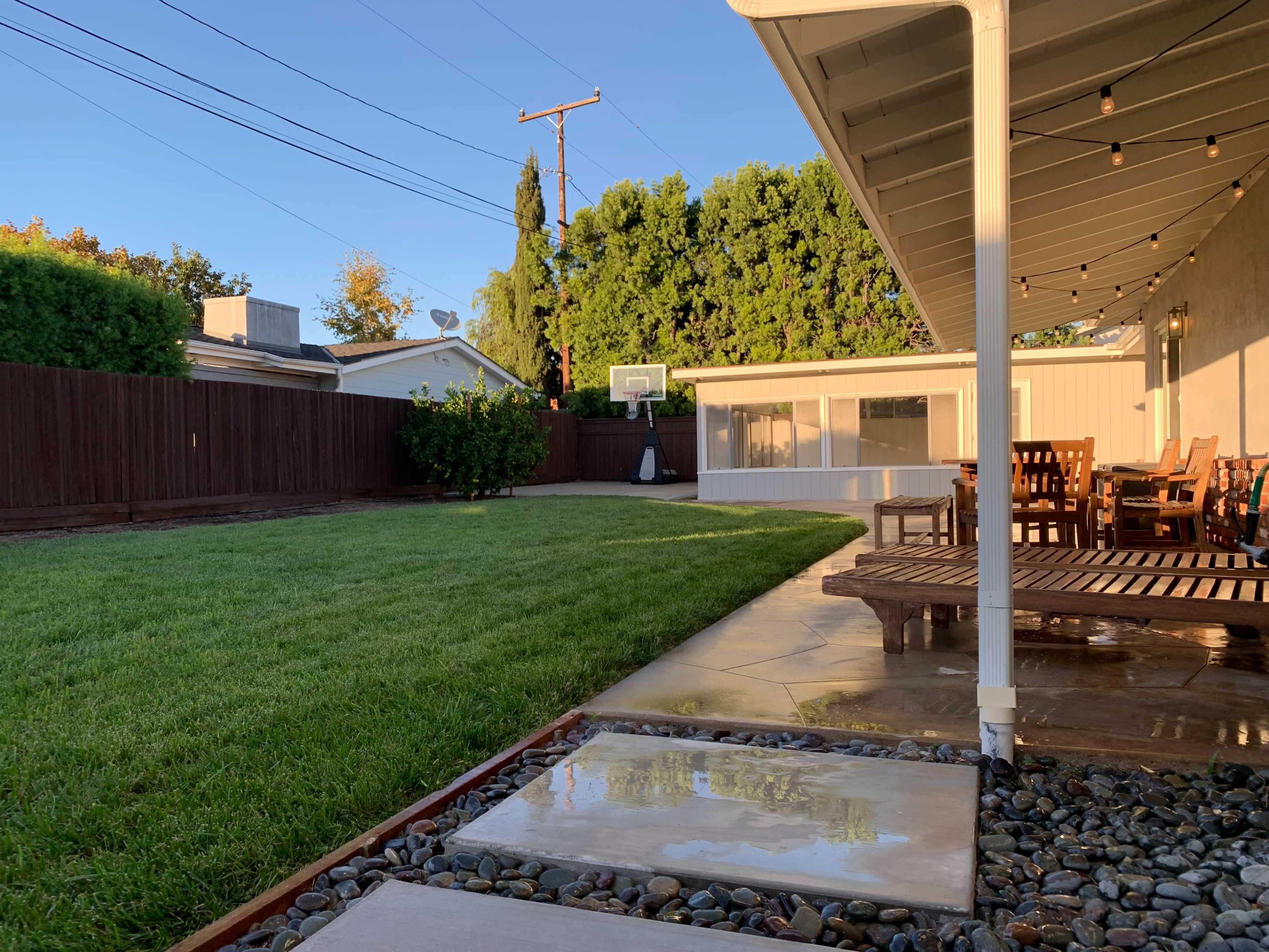 A backyard with a well-maintained lawn, a patio area with wooden furniture, and a basketball hoop in the distance.