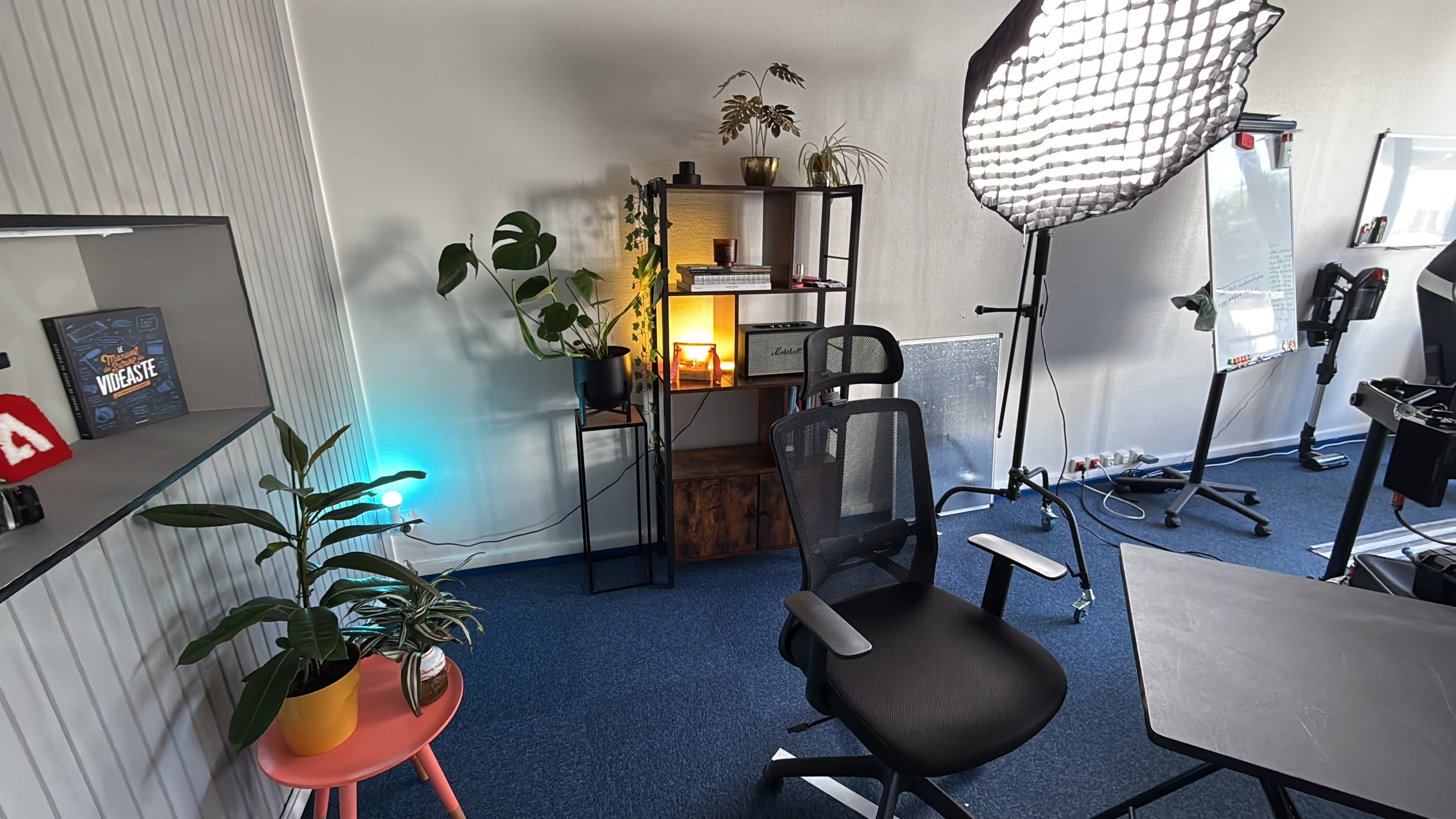 The image shows a well-lit workspace featuring a black office chair, a table, a bookshelf with plants and lighting, and a whiteboard in the background.