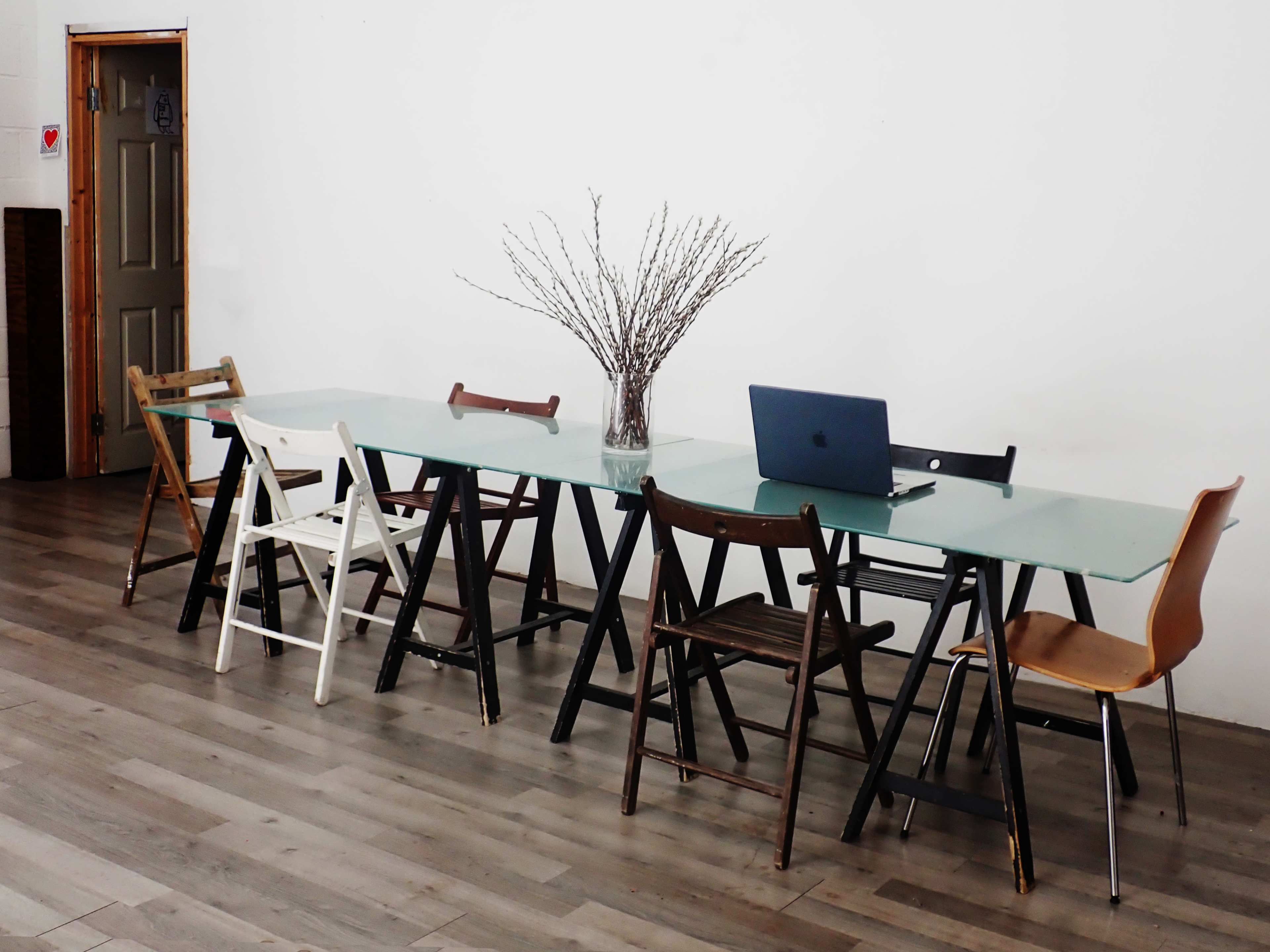 A long glass table with a laptop and a vase of branches is surrounded by various wooden chairs in a spacious room.