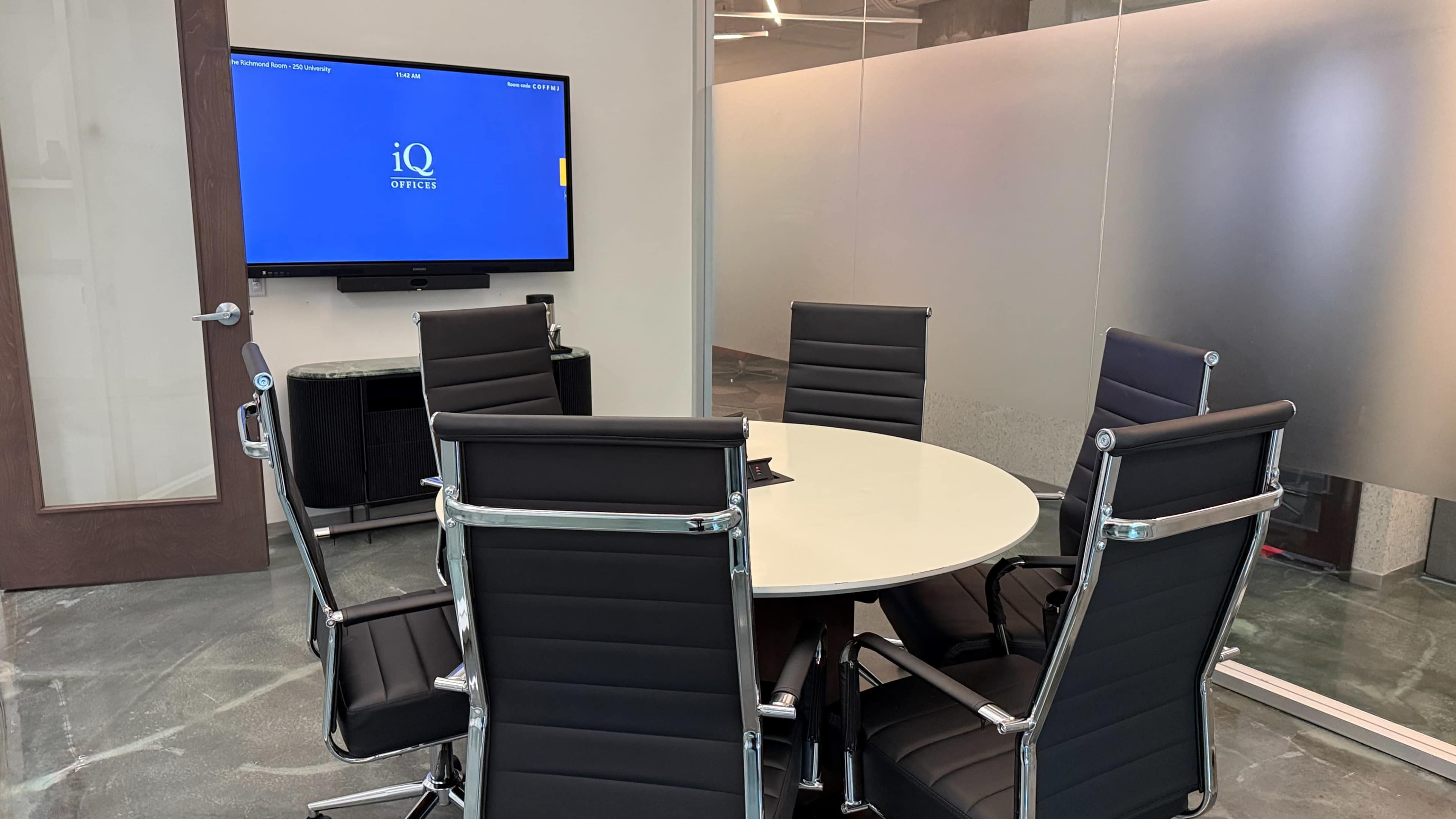 The image shows a modern conference room featuring a round table, six black leather chairs, and a wall-mounted television displaying the logo of a company.