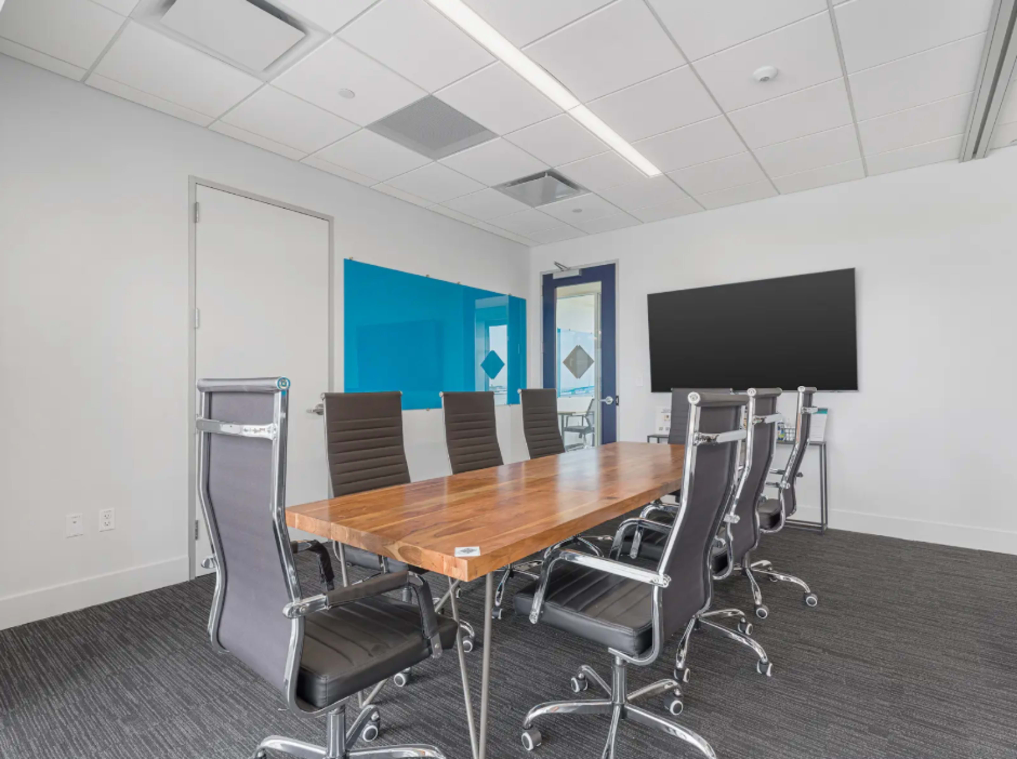 The image shows a modern conference room with a long wooden table surrounded by black leather chairs, featuring a blue whiteboard on one wall and a large screen on another.