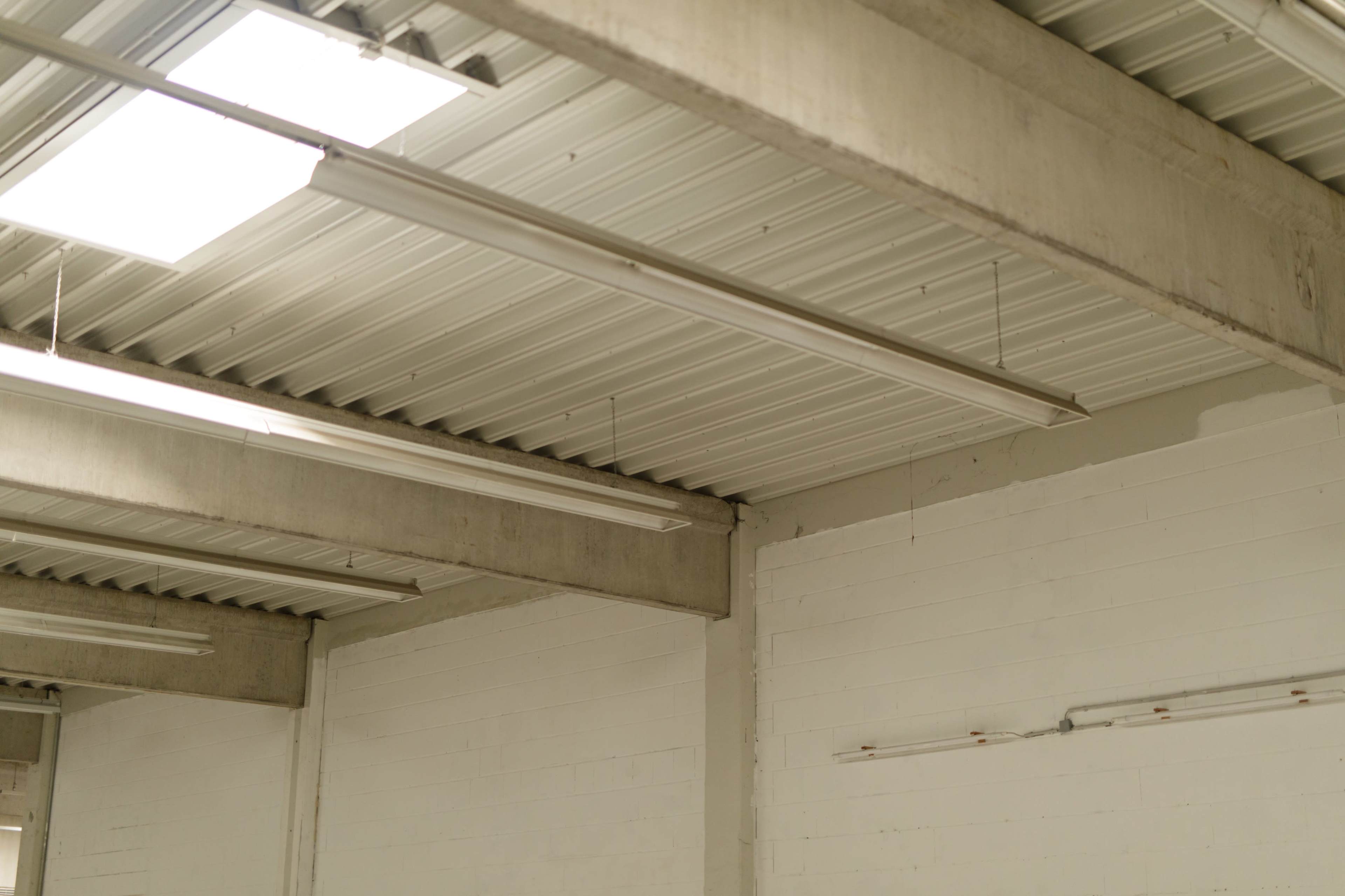 The image shows a section of an industrial space featuring a metal ceiling with fluorescent light fixtures and concrete support beams.