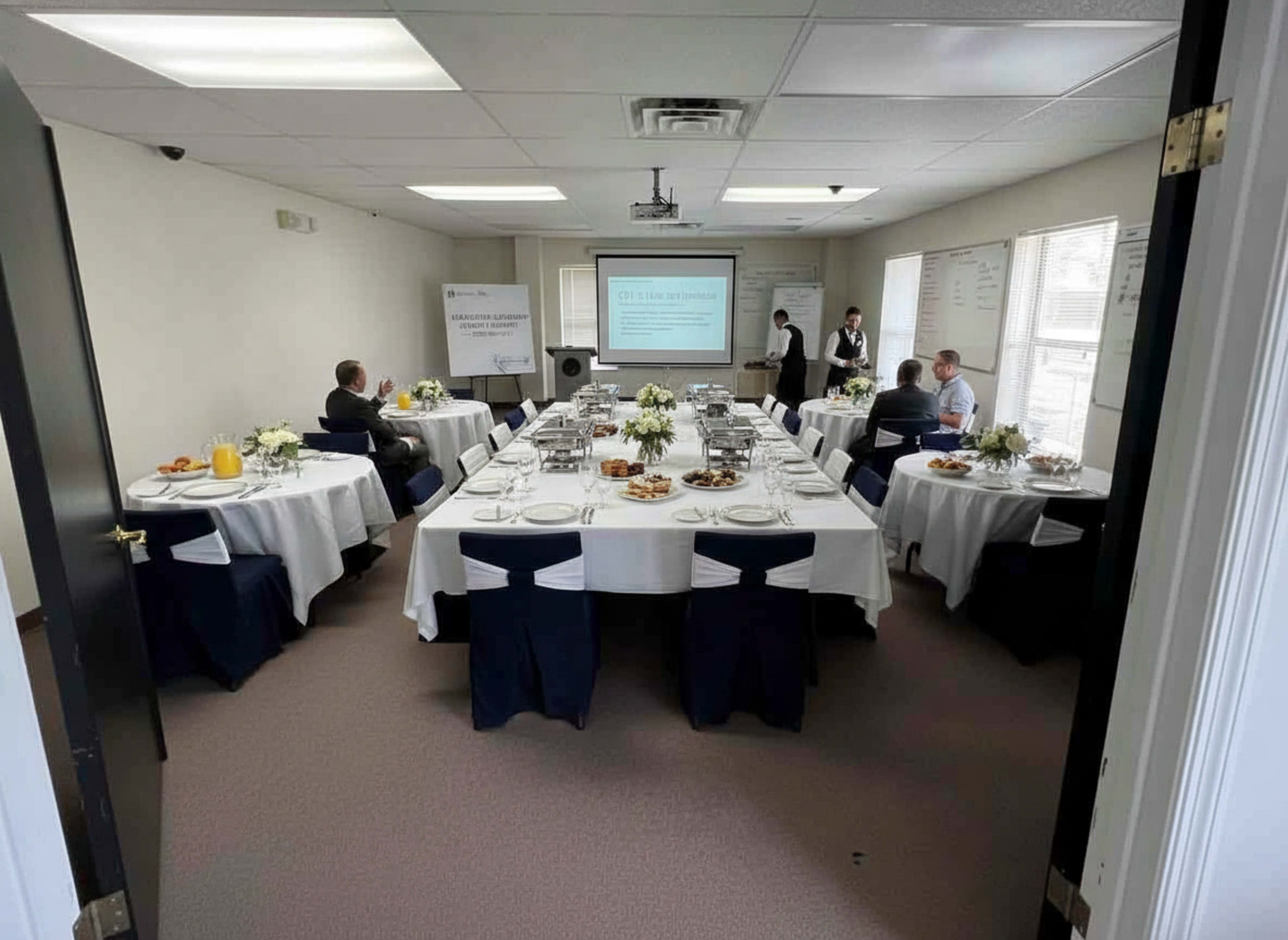 A conference room is set up with several round tables, each covered with white tablecloths and arranged with food, while a presentation screen displays information at the front.