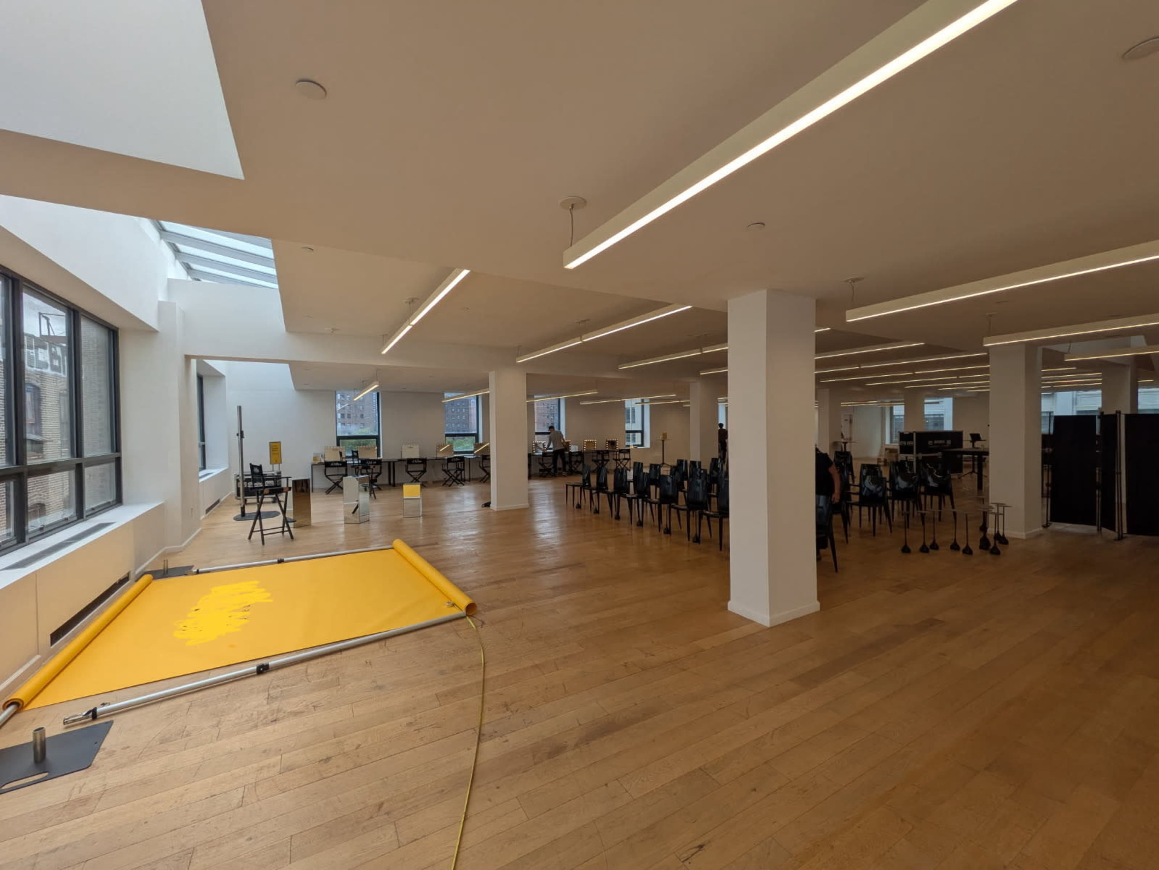 The image shows a spacious, well-lit room with wooden flooring, featuring chairs arranged in groups and a large yellow canvas on the floor.