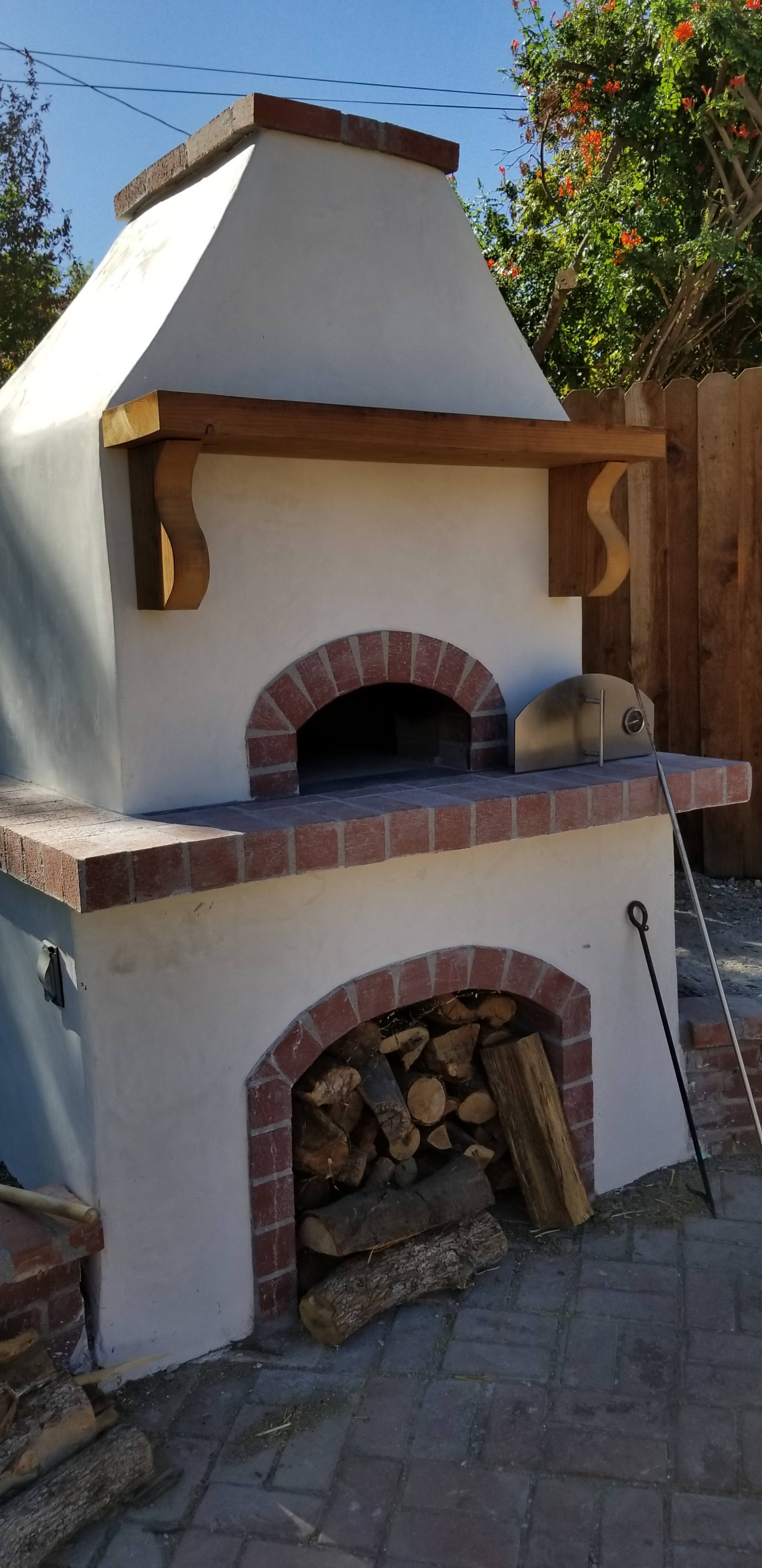 An outdoor pizza oven made of stone and brick, with a stack of firewood stored underneath.
