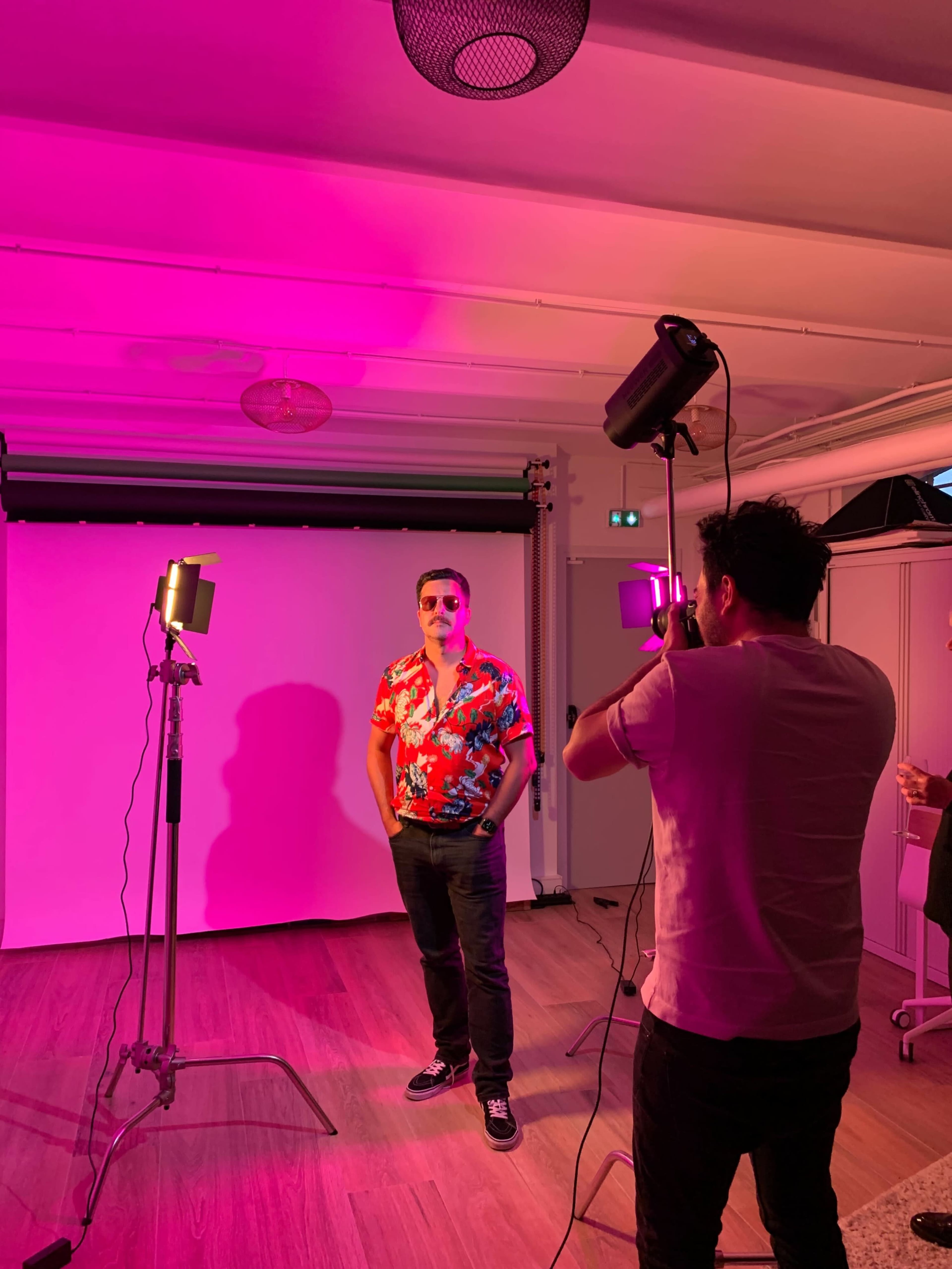 A man in a floral shirt poses for a photo while another person adjusts the camera under pink lighting.