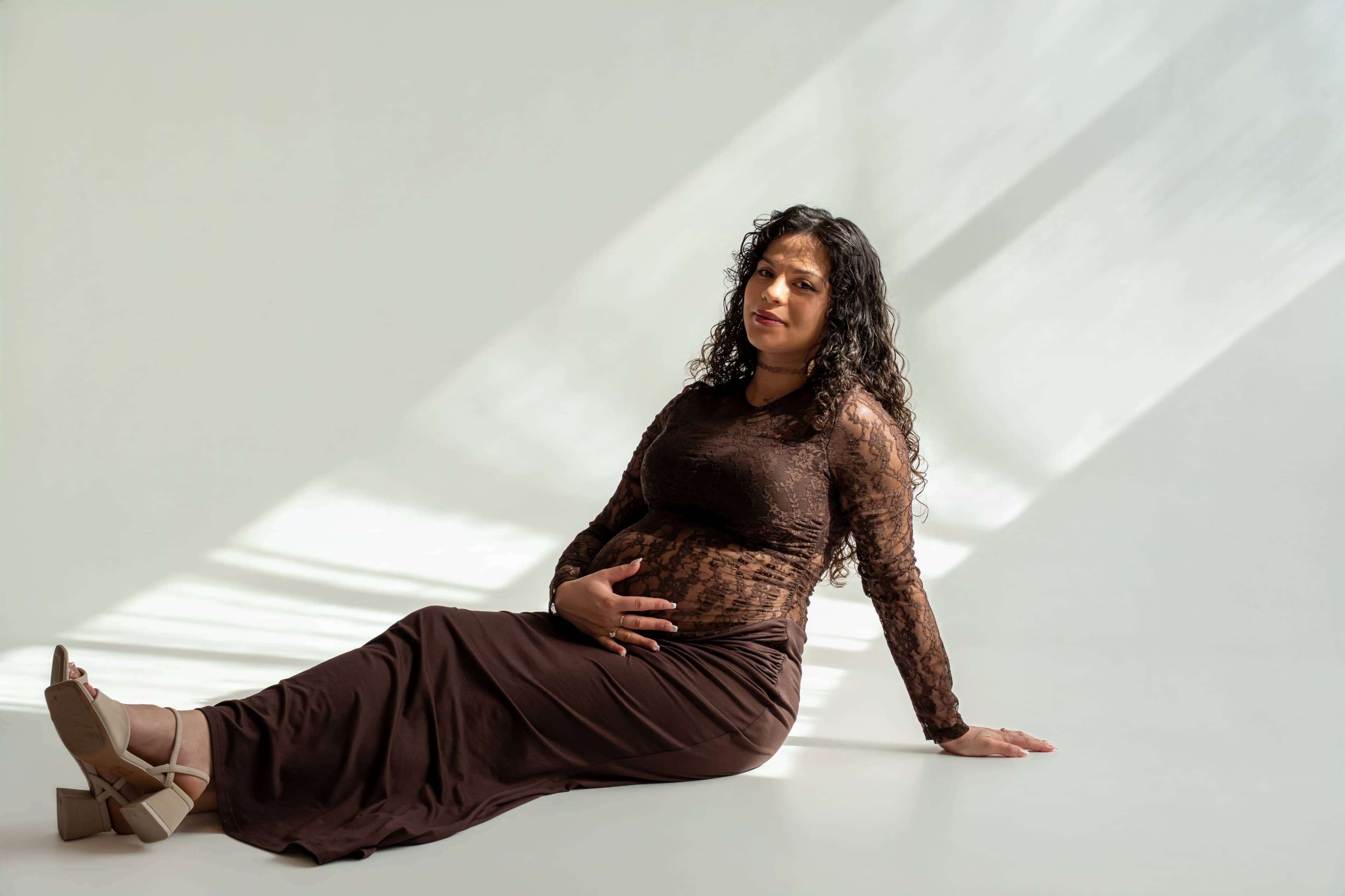 A woman in a brown dress sits on a white floor, gently cradling her pregnant belly while soft sunlight casts shadows around her.