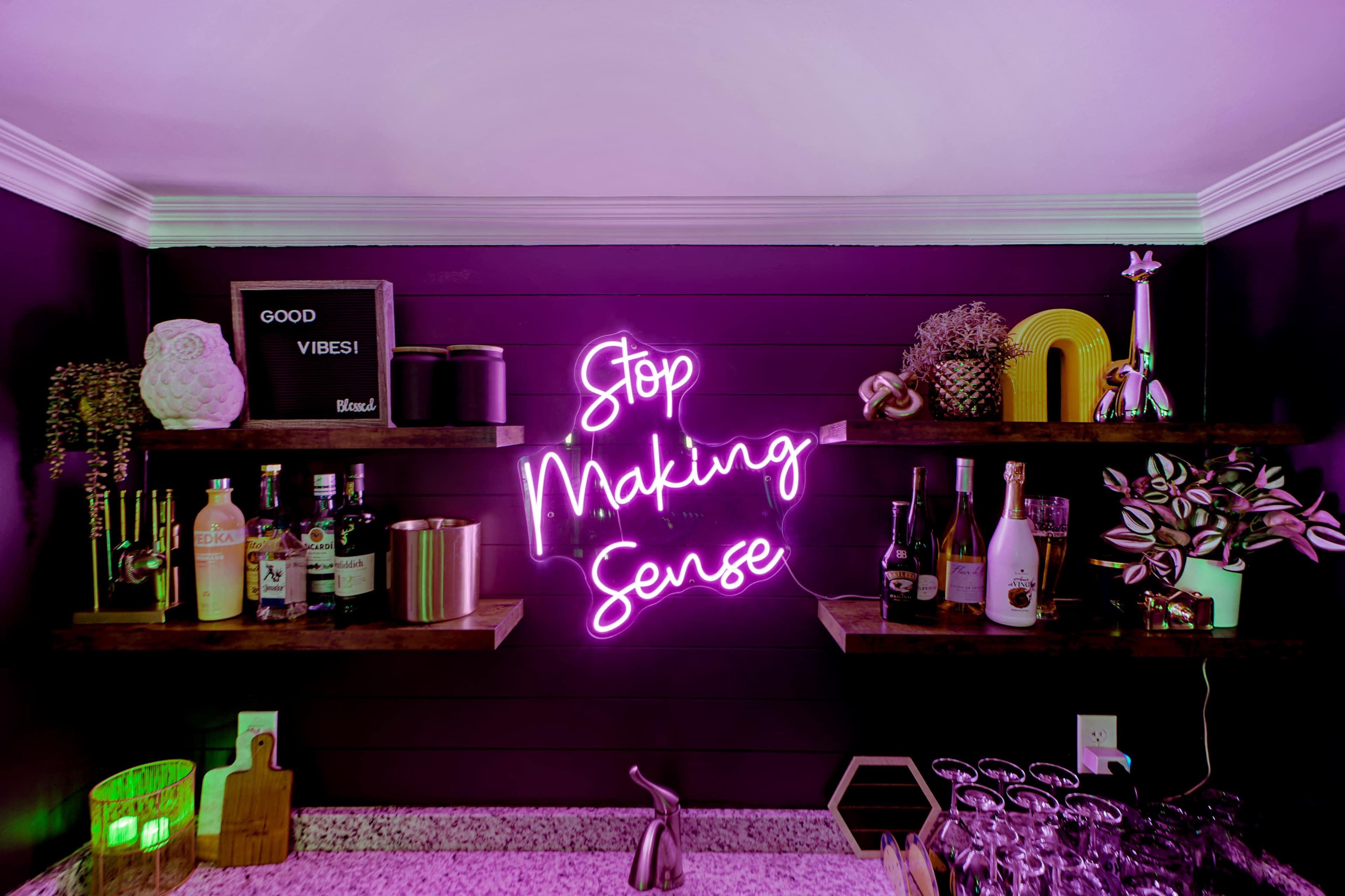The image shows a bar setup with neon signage that reads "Stop Making Sense" against a dark wall, complemented by shelves filled with various bottles and decorative items.