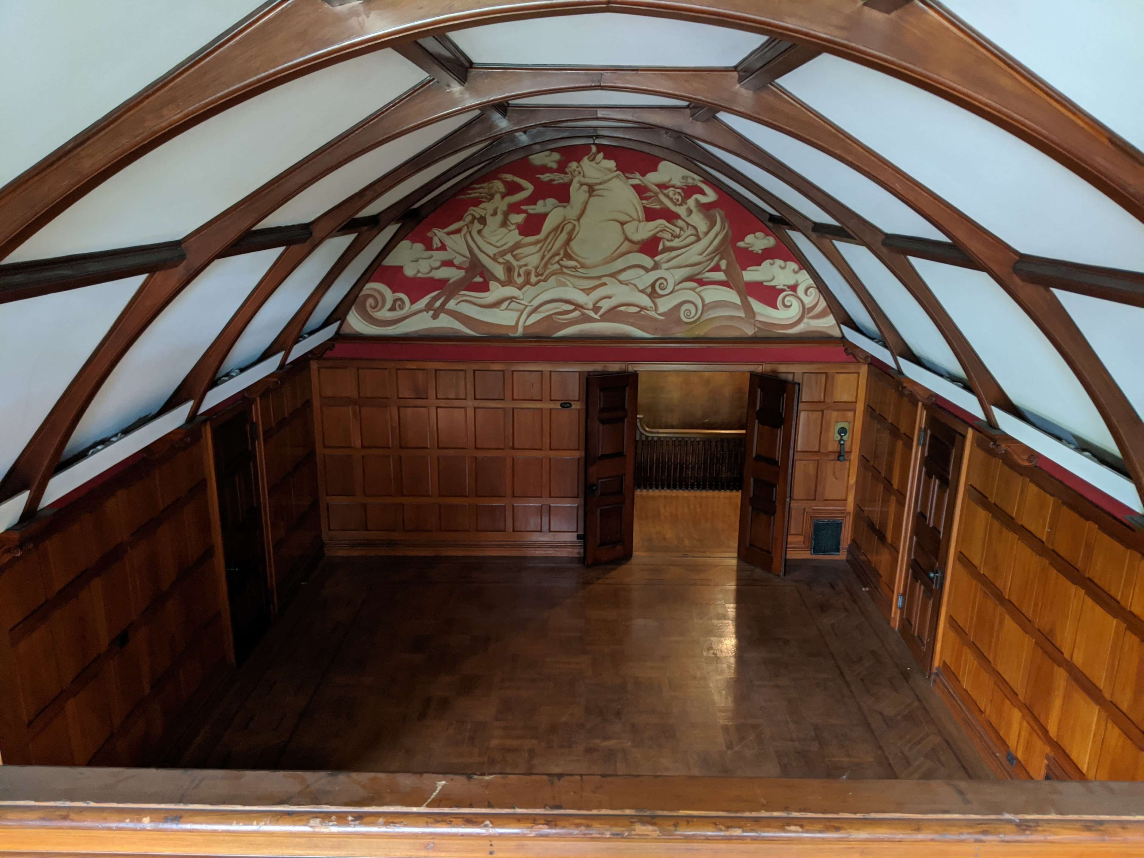 A wooden room with an arched ceiling features a mural of figures on a horse, and has two open doors leading to a darker space.