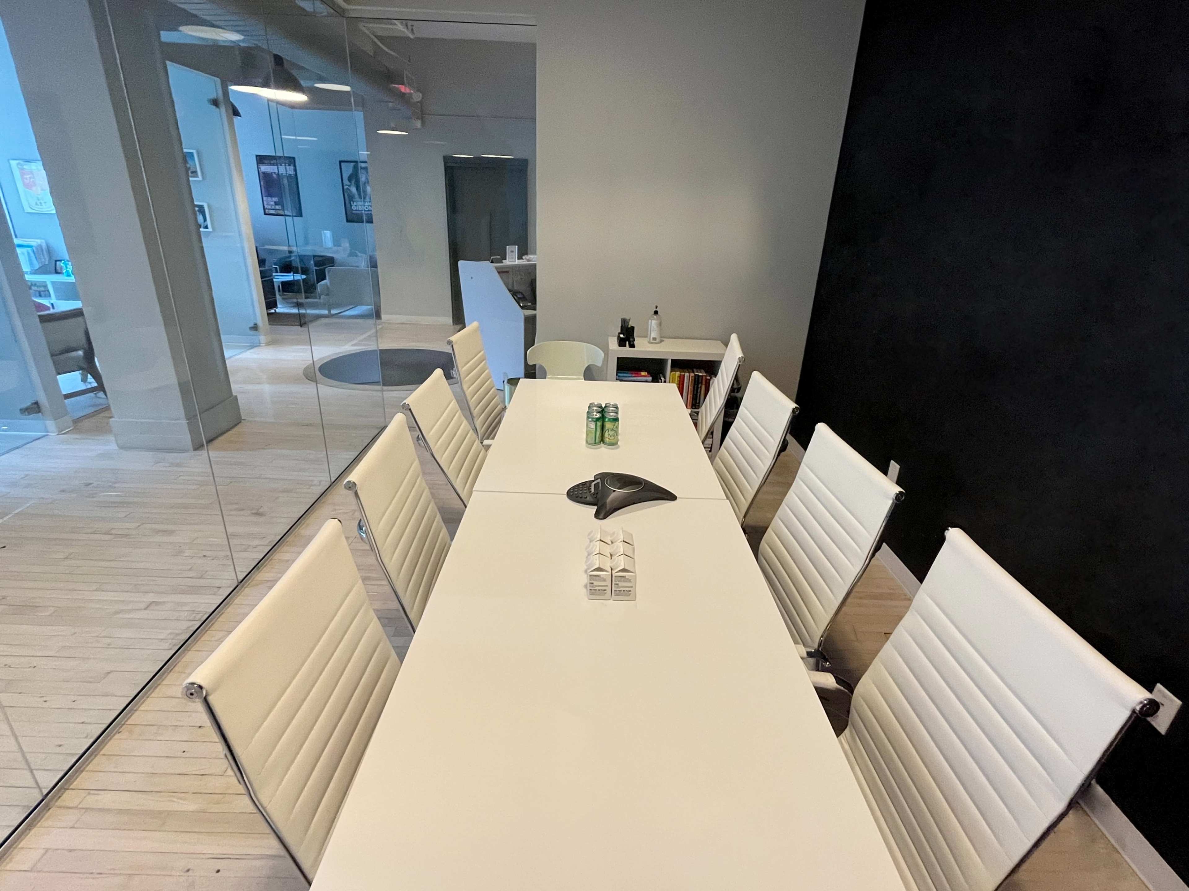 The image shows a minimalist conference room with a long white table surrounded by six white chairs, featuring a microphone and several glass containers in the center.