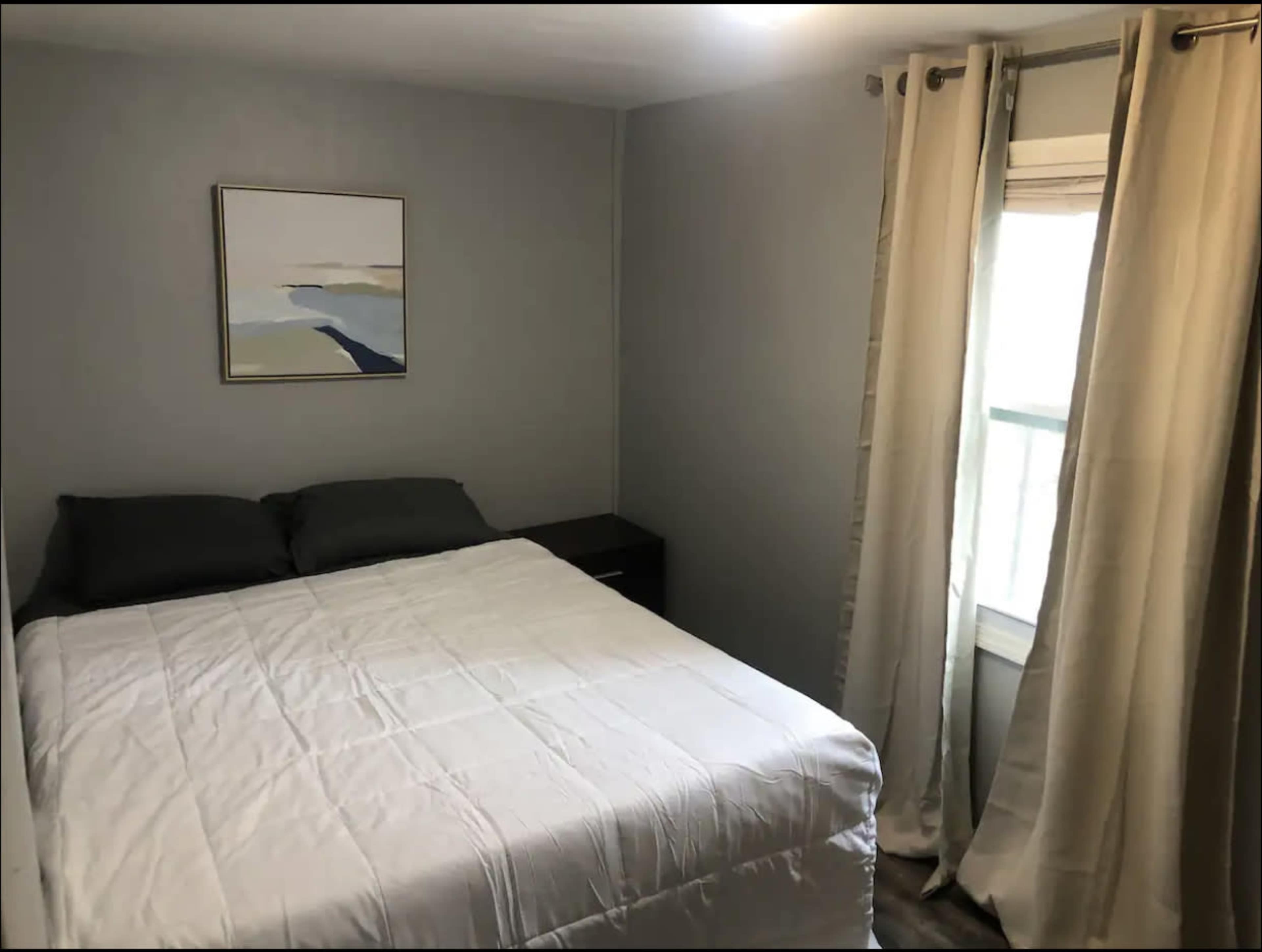A small bedroom features a bed with gray pillows, a nightstand, and a window covered by long curtains, along with a painting on the wall.