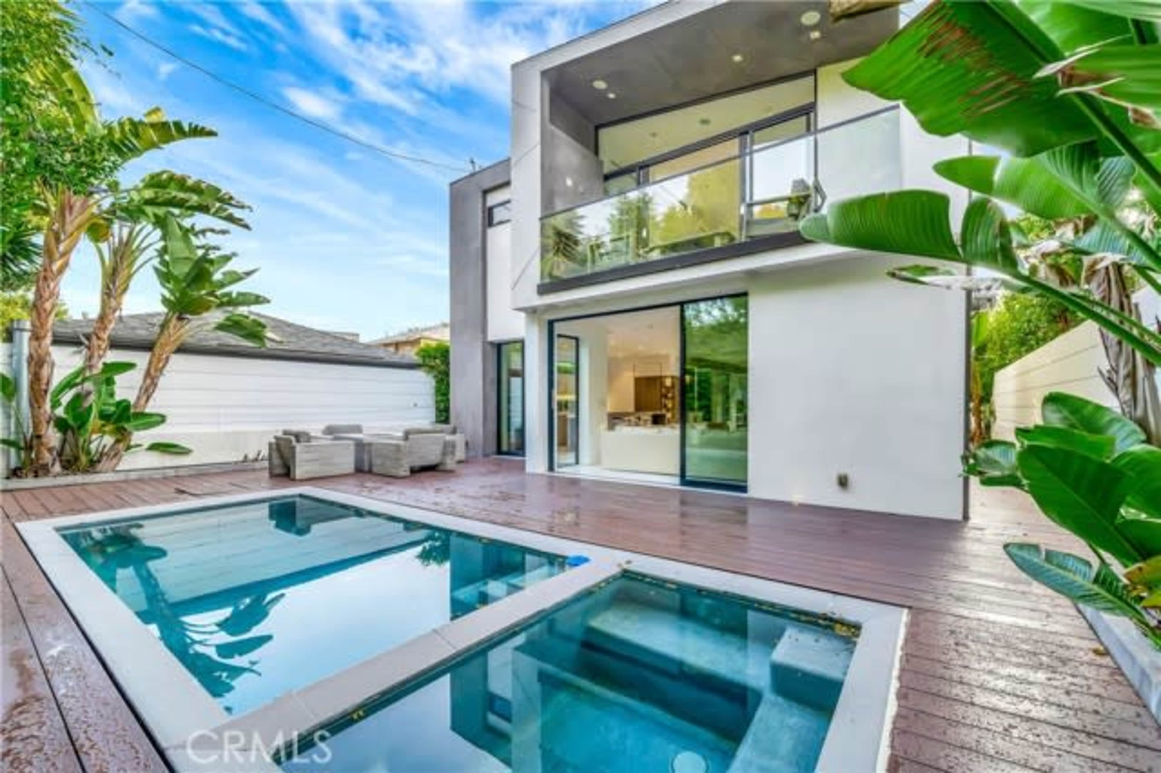 The image shows a modern two-story house with large glass doors opening to a wooden deck and a small swimming pool surrounded by tropical plants.