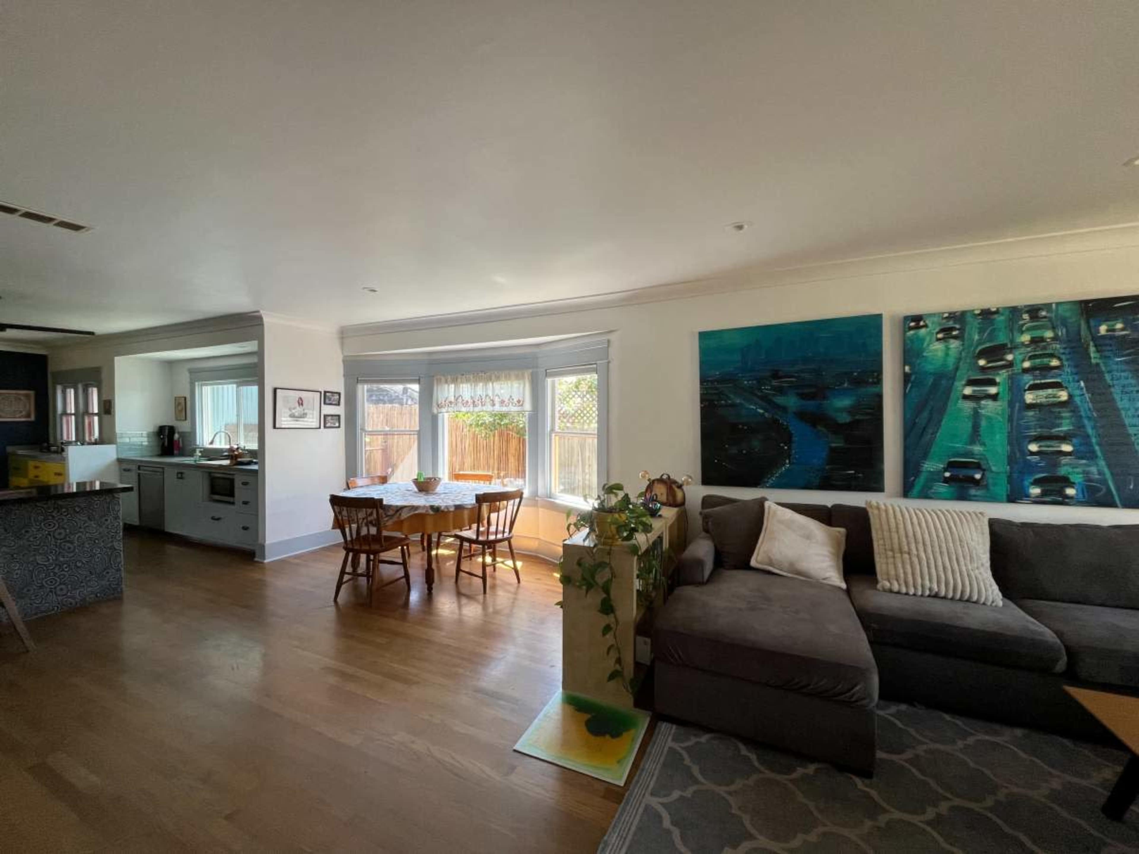 A spacious living area features a gray sectional sofa, a dining table, and a kitchen visible in the background, illuminated by natural light through large windows.