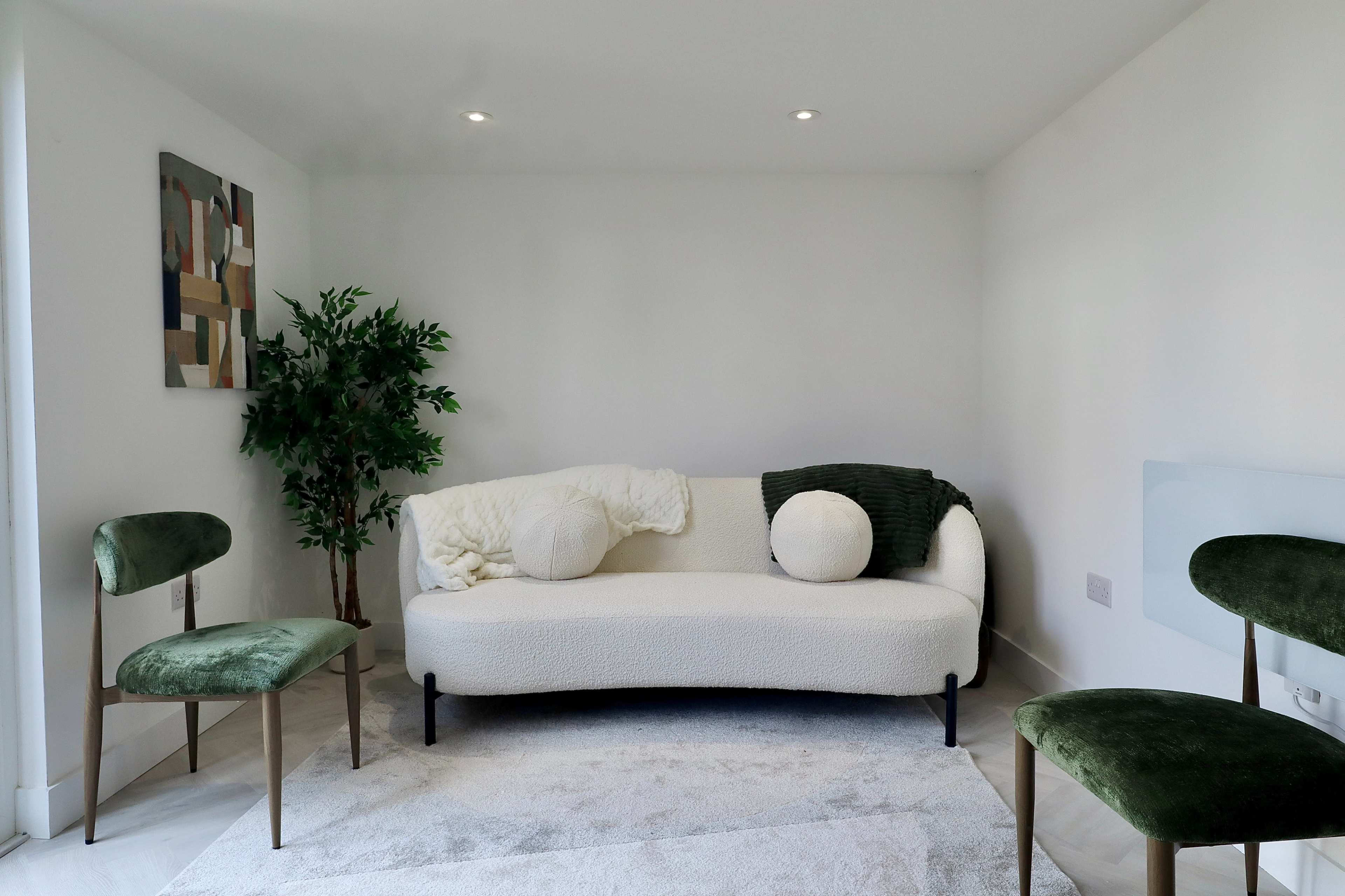 The image features a minimalist living space with a cream-colored couch, two green chairs, a small indoor plant, and a light-colored rug on the floor.
